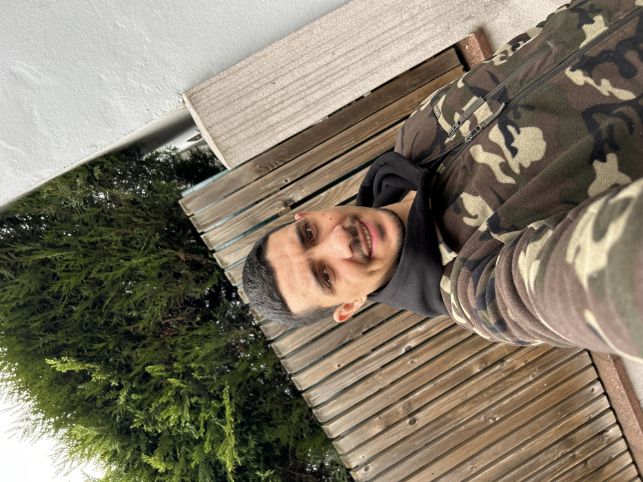 An outdoor portrait in Schiltigheim, France, captures an adult male in a selfie. The man, positioned centrally, possesses short dark hair, dark eyes, a mustache, and a short beard, looking directly at the camera with a slight smile. He wears a camouflage-patterned fleece jacket, featuring brown, green, and beige hues, with a central zipper and two zippered chest pockets. A black hooded sweatshirt is visible beneath the jacket. His right arm is visible in the lower right foreground, consistent with holding a camera. The background on the left features a vertical slatted wooden fence, behind which dense green coniferous foliage is visible. To the right of the subject, a light grey wall meets a textured, light-colored concrete or stone slab. A narrow strip of reddish-brown ground is visible at the bottom edge of the frame.