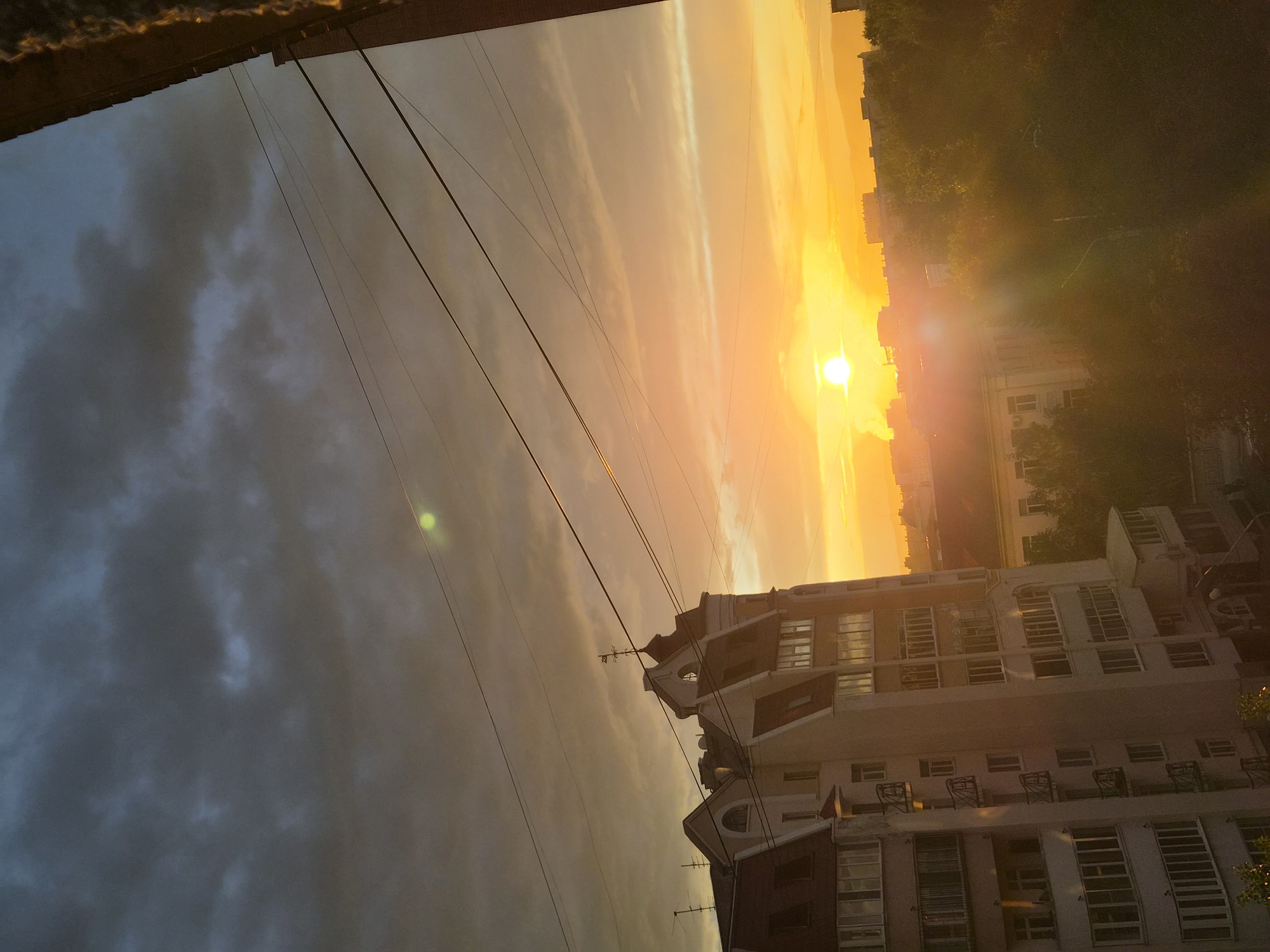 This is a photograph taken at sunset in the city of Chita, Russia. The sky is filled with dramatic clouds, ranging from dark grey to vibrant orange and yellow hues. The sun is low on the horizon, casting a warm glow across the scene and creating lens flare. In the foreground, the upper floors of a multi-story building with a distinctive mansard roof and dormer windows are visible. The building is light-colored with red-tiled roofs and dark-trimmed windows. Several electrical wires run diagonally across the sky. In the middle ground, other buildings and trees are silhouetted against the bright sky. The overall impression is of a city at dusk, with the warm light of the setting sun illuminating the urban landscape.