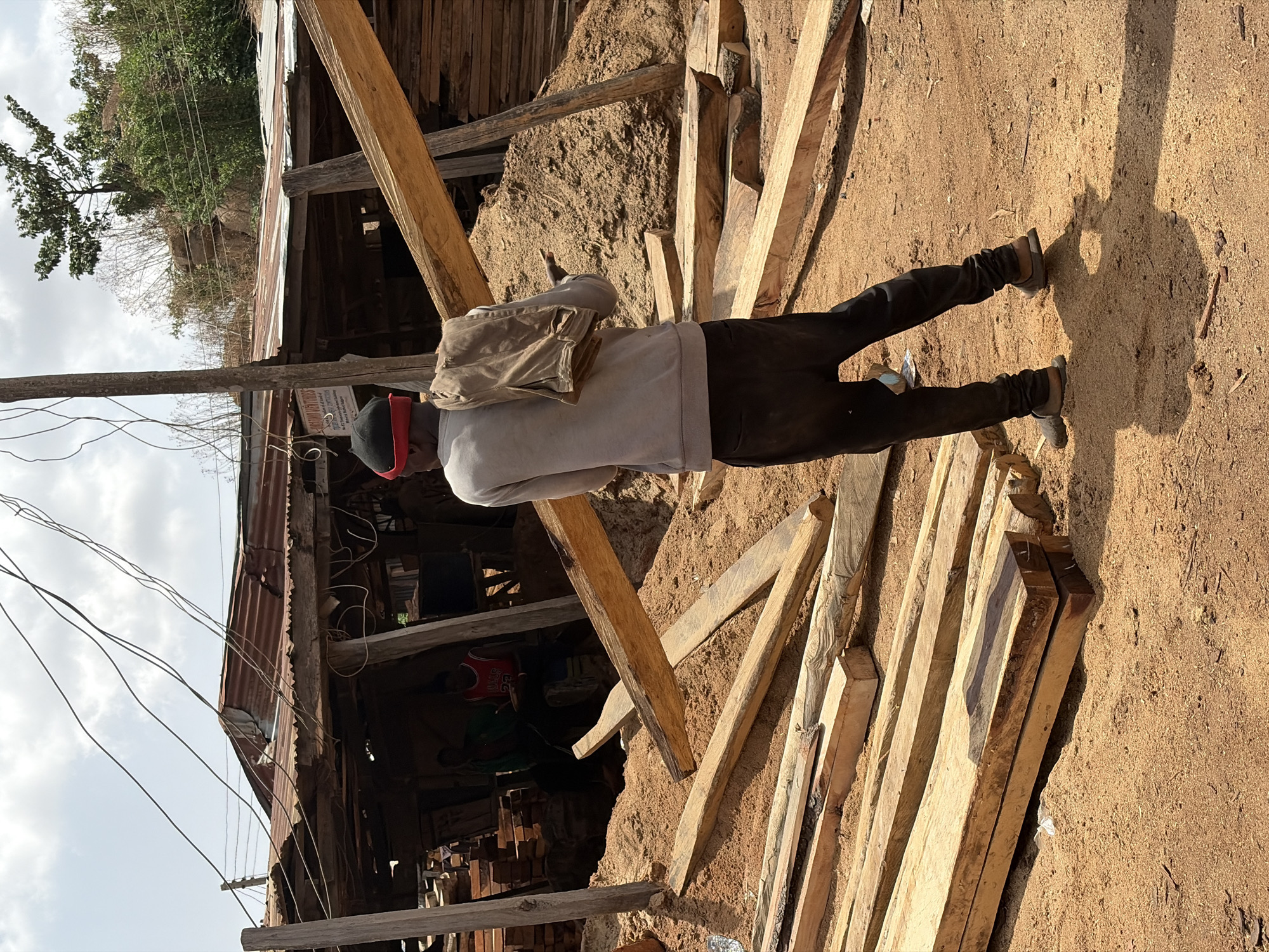 A man stands amidst scattered wooden planks and sawdust-like material on a dirt ground. He wears dark trousers, brown shoes, a light grey sweatshirt, and a dark hat with a red accent. He has a beige-colored bag slung over his shoulder. To his left, a partially constructed wooden structure with a rusty corrugated metal roof is visible. Inside the structure, there are glimpses of other individuals and stacked materials. Overhead, utility wires stretch across the sky. The location is identifiable by the presence of the construction materials and rudimentary structure. This is Jos, Nigeria.