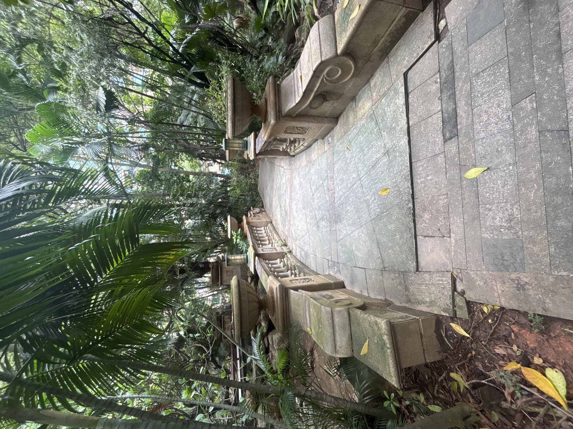 The image shows a paved pathway curving through a lush, green garden environment. The pathway is made of grey stone tiles and is bordered by a decorative stone railing with planters on top. In the background, dense tropical foliage, including palm trees and other greenery, creates a natural canopy, with glimpses of buildings visible through the leaves in the distance. Fallen yellow leaves are scattered on the pathway and surrounding ground, suggesting an outdoor setting. The style of the architecture and landscaping suggests a park or botanical garden.