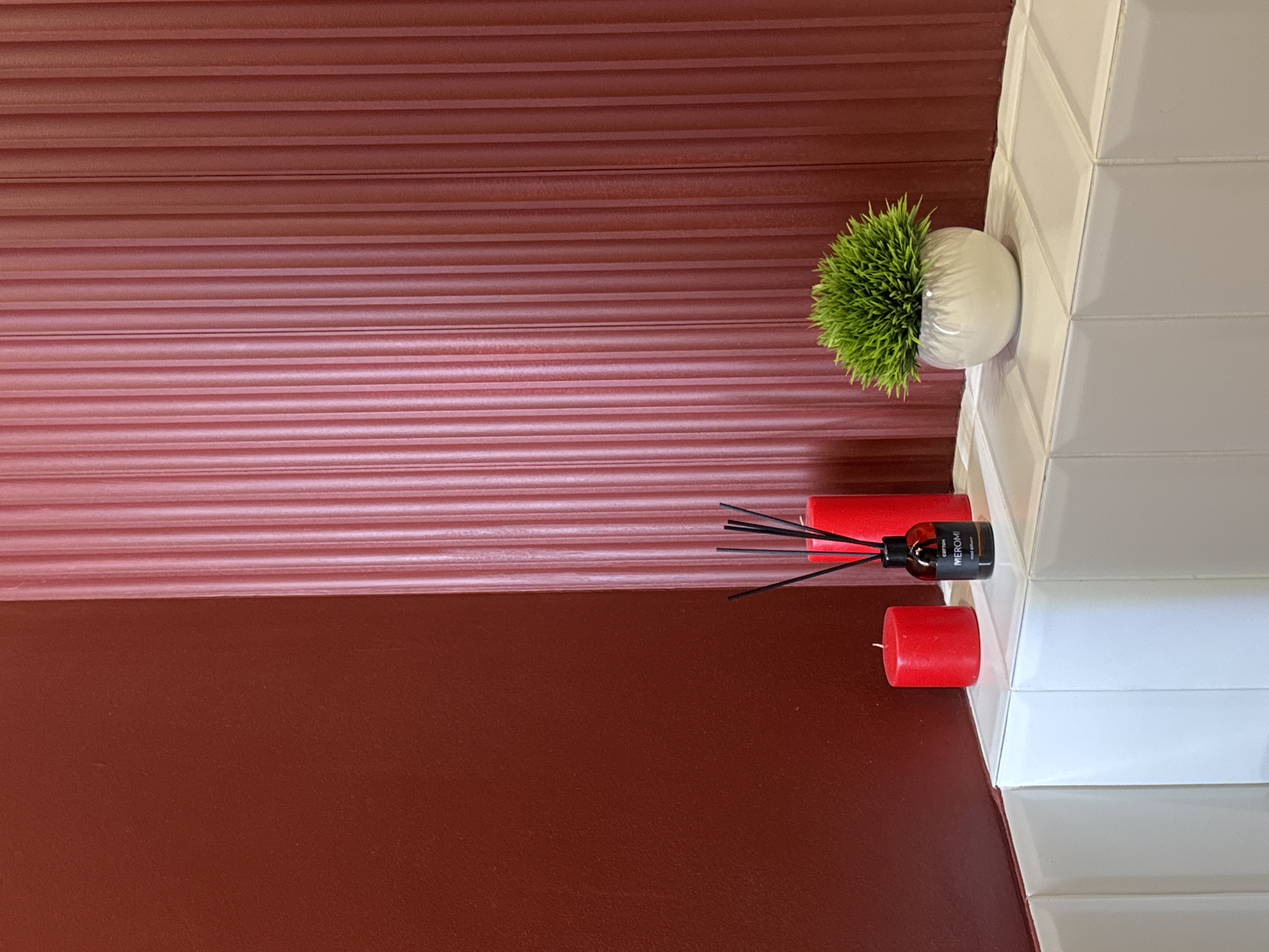 The media file displays a close-up, vertically oriented shot of a decorative arrangement on a surface. The background features a textured red wall with horizontal lines. To the right, a white tiled surface with a geometric pattern is visible.
On the tiled surface, a small, round, white pot contains a bright green, spiky artificial plant. Next to the plant, a reed diffuser with a dark glass bottle and black reeds is placed. The bottle has a label with the text "MEROMI" and "COTTON" printed on it. Beside the diffuser are two red cylindrical candles.
The lighting appears to be indoor, with the light source illuminating the objects from the right. The overall scene suggests a decorative corner in a room. There are no people, and no specific time of day or weather conditions can be inferred. The location context provided is Патрушева, Russia, which is not visually evident from the image itself.