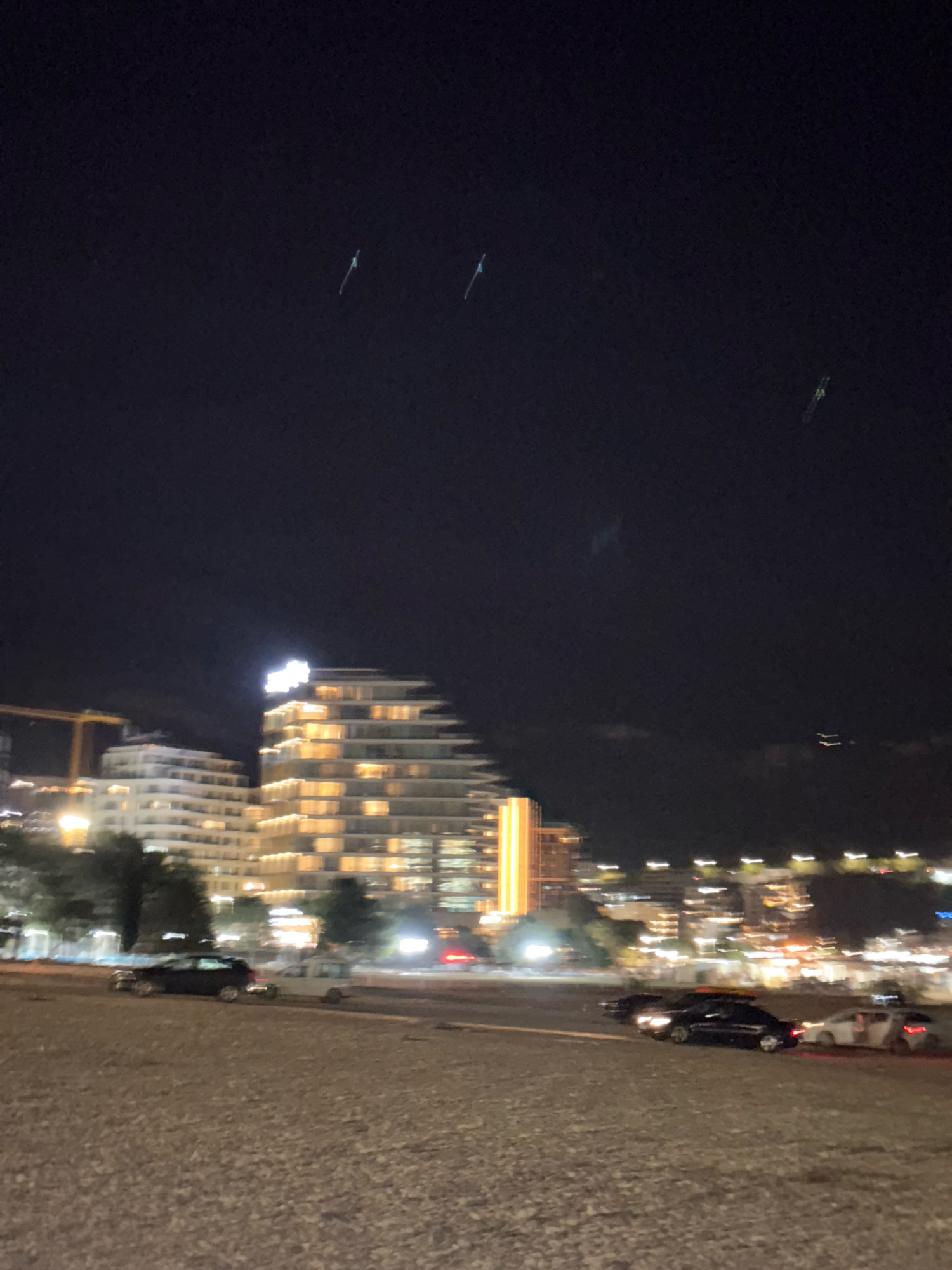 In the city of Boreti, Montenegro, at night, a modern, stepped building illuminated by warm lights is visible. In front of it, a series of lower buildings also glow with interior lighting. A construction crane stands to the left of these buildings. Several cars are parked or driving on a road in the foreground, their headlights and taillights creating streaks of light. The sky is dark with a few faint lights that may be aircraft or distant lights. The ground in the foreground appears to be gravel or a similar textured surface.