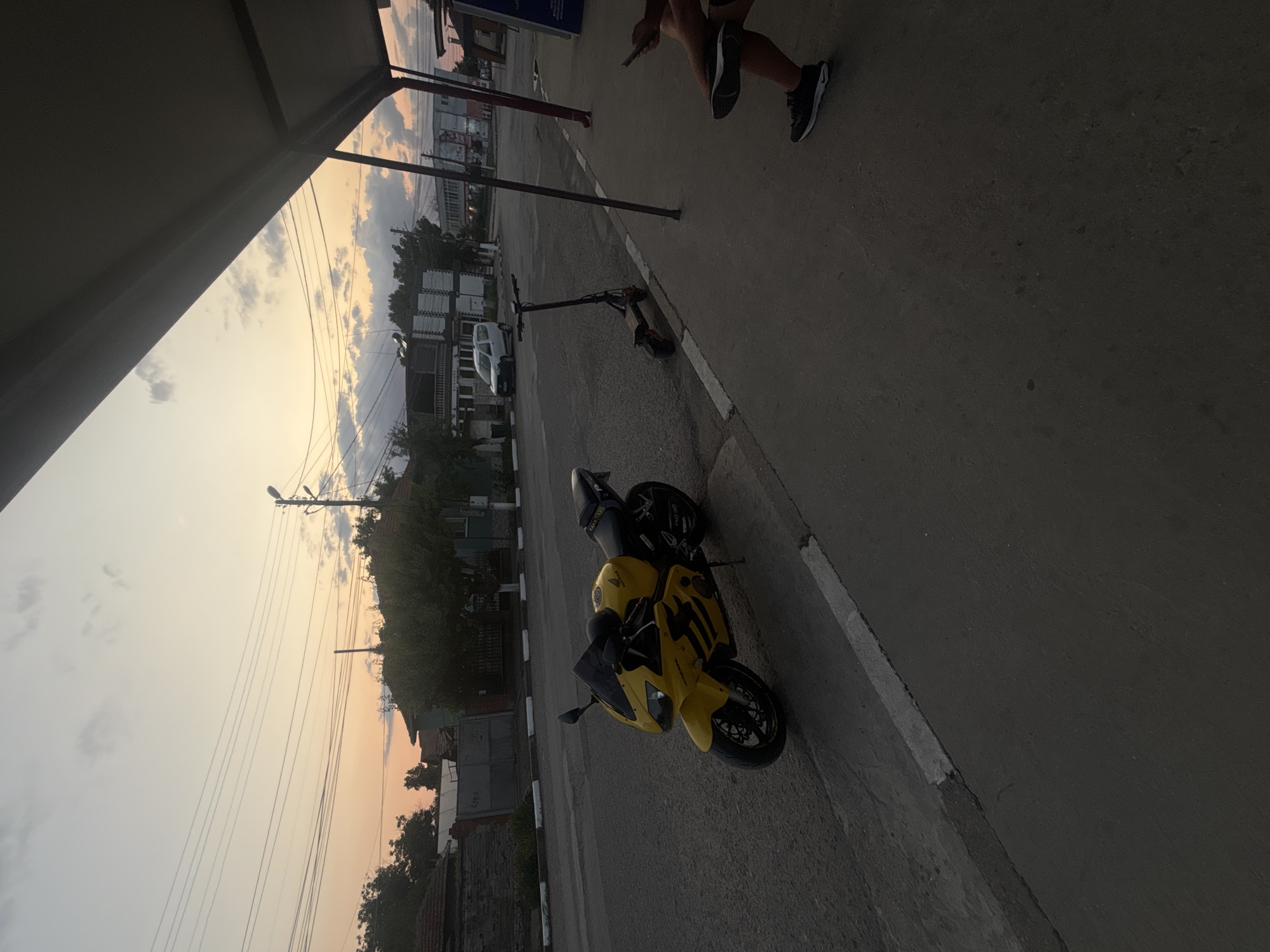 A yellow Honda CBR motorcycle is parked on the side of a street in Marten, Bulgaria, next to a sidewalk with a partial view of a bus stop shelter. An electric scooter is parked behind the motorcycle. In the background, there are buildings and trees under a twilight sky with visible power lines. A person is seated on the sidewalk on the right side of the image, with their legs visible.