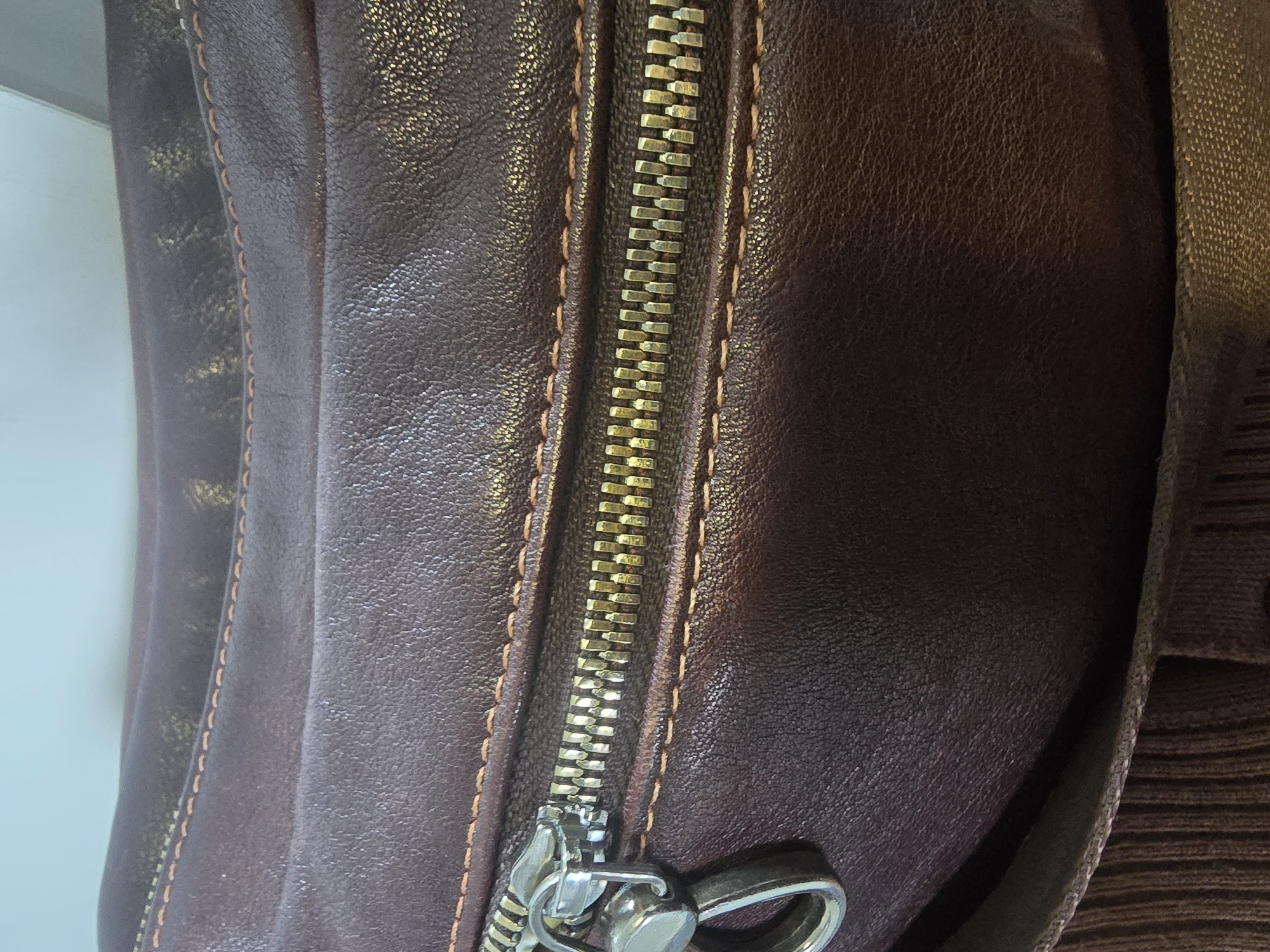 The image is a close-up, high-angle shot of a dark brown leather bag with a silver zipper. The leather has a textured, slightly worn appearance. A thick, tan-colored strap is visible at the bottom of the image, with a ribbed brown fabric beneath it. The background is a blurry, light blue, suggesting an indoor setting or an overcast sky. There are no people or identifiable landmarks in the image. The provided crops highlight different sections of the bag, focusing on the texture of the leather, the metallic sheen of the zipper teeth, and the stitching details. The scene is devoid of any context that would place it in Ulan-Ude, Russia, or any other specific city.