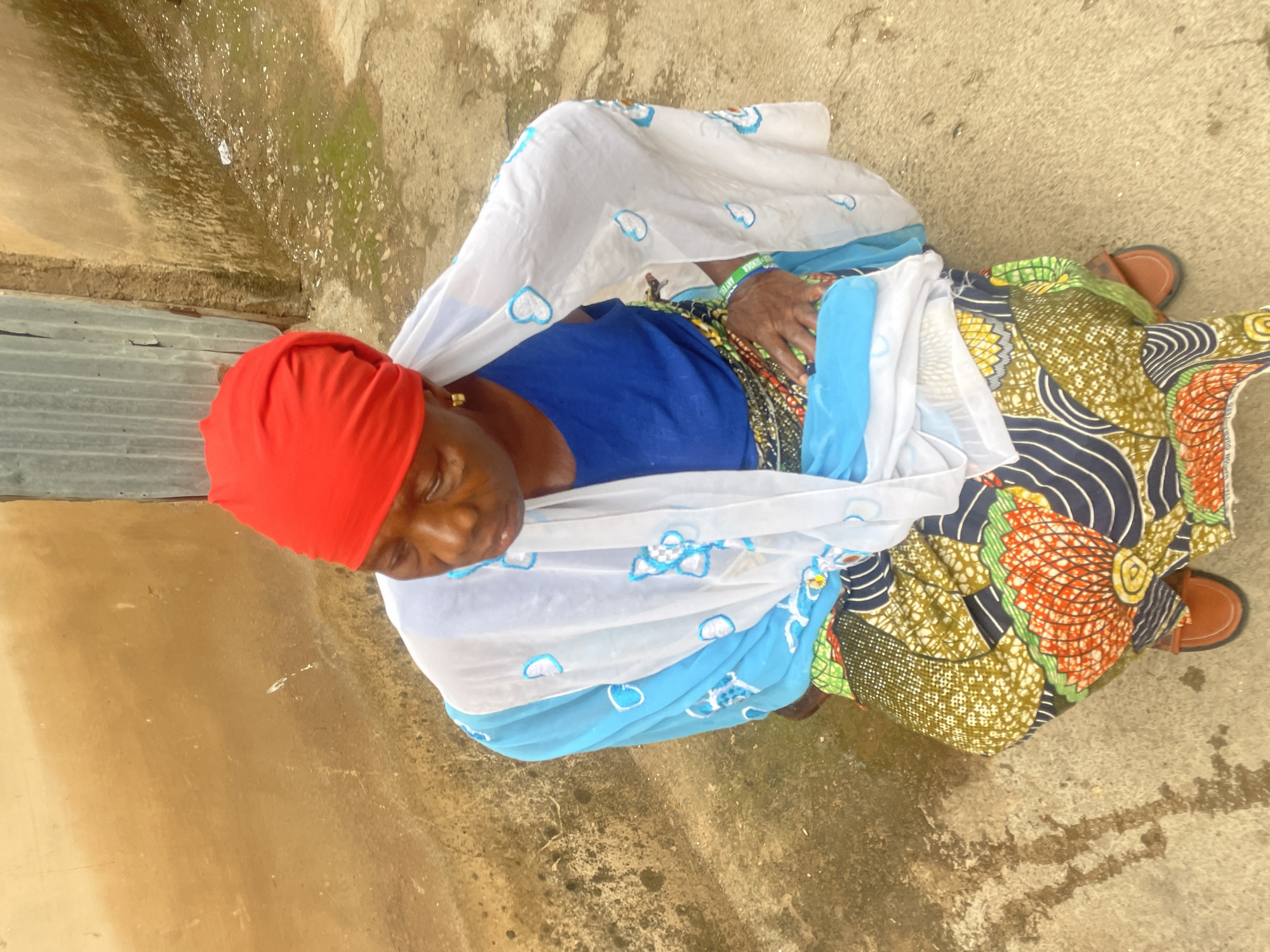 A high-angle, full-body shot depicts an African woman seated on a concrete surface outdoors. The location is Jos, Nigeria. The woman wears a red head covering, a blue top, and a patterned, colorful skirt. A white shawl with blue floral prints is draped over her. She is wearing brown sandals. The concrete surface appears weathered. A wall is visible on the left side of the frame.