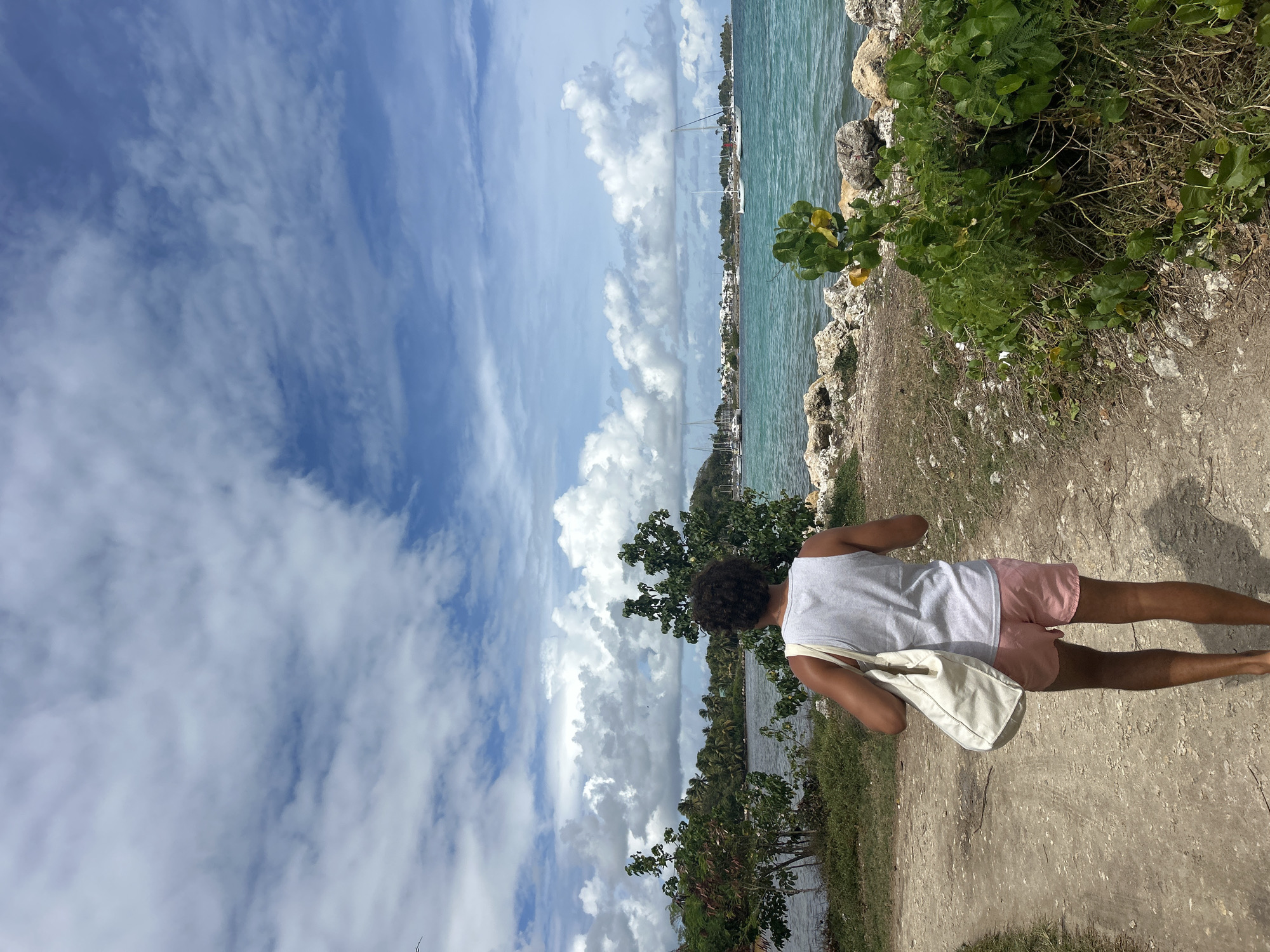 A medium shot shows a person walking on a dirt path by the water. The person, seen from the back, is wearing a white tank top and pink shorts, and carrying a white canvas bag on their shoulder. To the left, beyond some trees and shrubs, is a body of water, followed by a sky filled with puffy white clouds. To the right of the path, vegetation grows to the edge of the water, where there are some rocks and a clear view of boats. In the distance are some buildings. This scene is in Sainte-Anne, France.