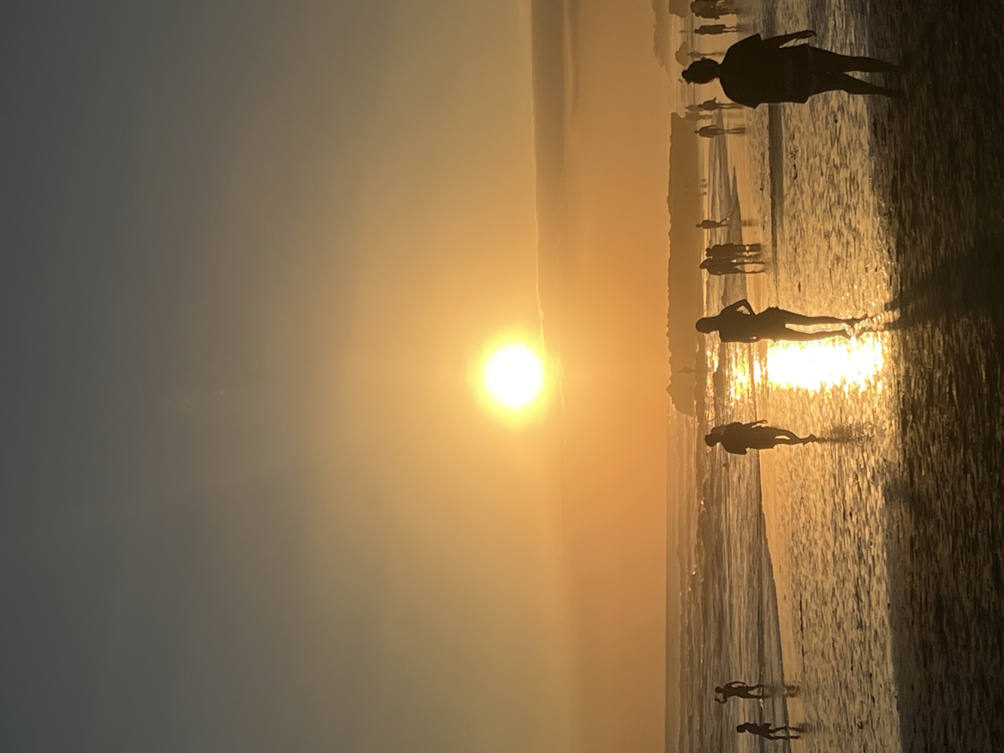 A photograph captures a sunset on a beach in North Kuta, Indonesia. The sun, a bright orb, hangs low in the sky, casting a warm golden glow that reflects off the wet sand and gentle waves. Silhouettes of people are scattered across the beach, some walking along the shoreline, others wading in the water, and a few standing further up the sand. The figures are indistinct due to the low light conditions. The ocean stretches out to the horizon, with small waves lapping the shore. The sky transitions from a pale blue at the top to a vibrant orange and yellow near the horizon. The overall scene conveys a peaceful and serene atmosphere as the day comes to a close.