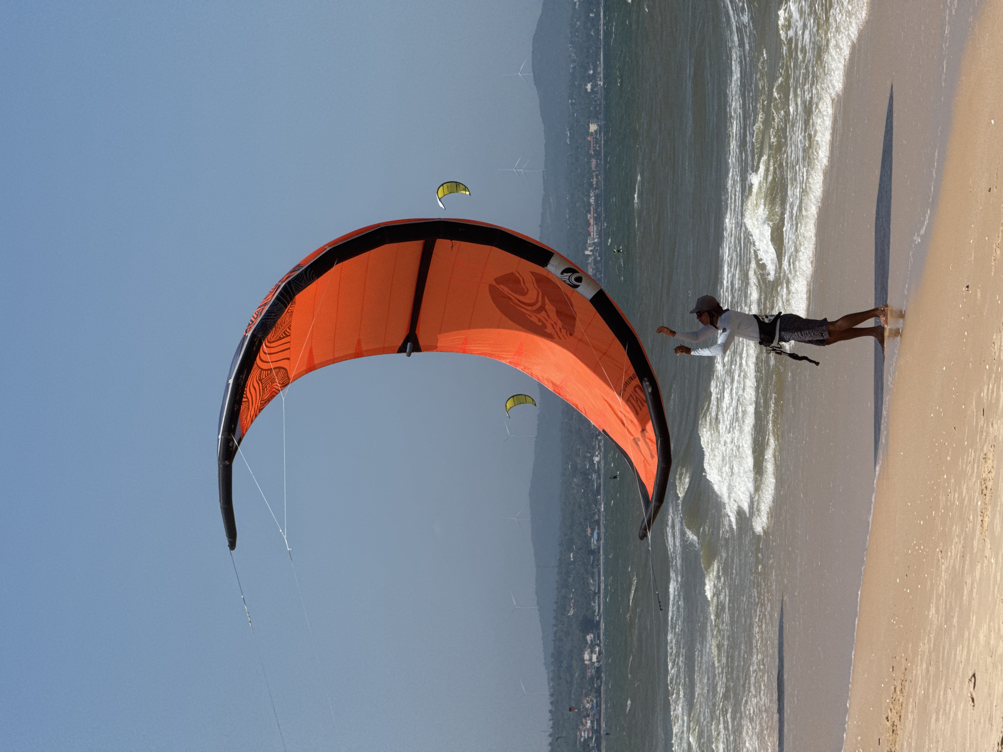 A person stands on a sandy beach in Mũi Né, Vietnam, holding the lines of a large, orange kite. The kite is curved in a crescent shape, dominating the upper portion of the frame. The person is wearing a light-colored long-sleeved shirt, dark shorts, and a cap. Their arms are raised, holding onto the kite's control bar. The ocean behind them has choppy waves, and further in the distance, a hazy coastline with buildings and wind turbines is visible. Other smaller kites can be seen in the sky and on the water, indicating a popular spot for kitesurfing. The sky is clear and blue.