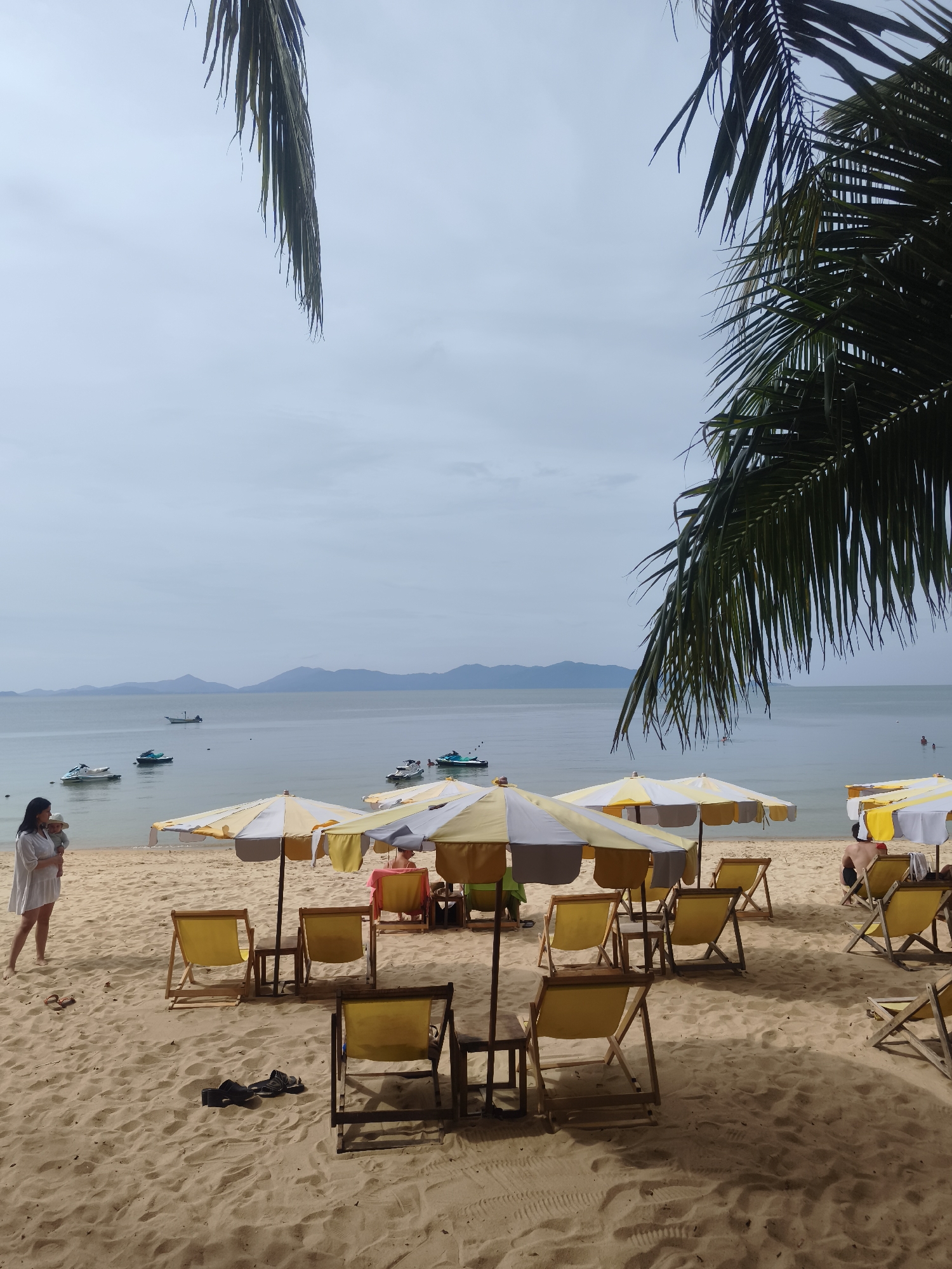 The photograph depicts a beach scene in Baan Mae Nam, Thailand. The beach has light-colored sand, on which a number of yellow and white striped beach umbrellas and corresponding beach chairs are arranged. A woman stands near the edge of the water, holding a child. Several motorboats are visible on the water in the distance. Palm trees hang down from the top corners of the frame.