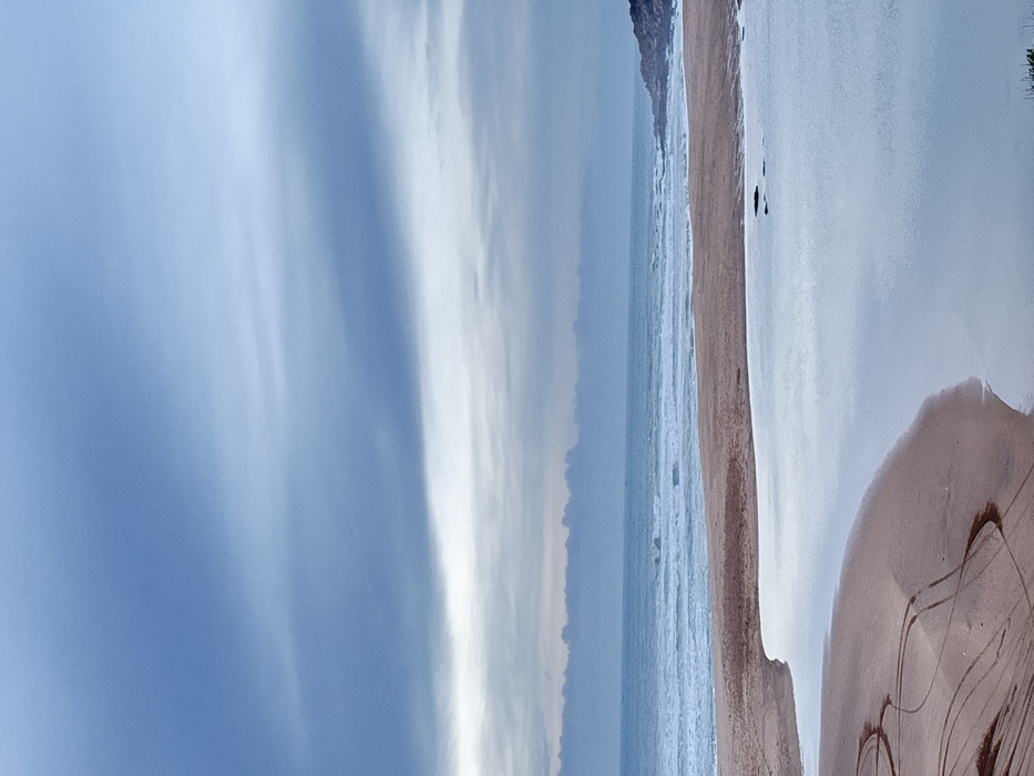 A landscape photograph presents a vast beach under a predominantly cloudy sky. The sky, encompassing roughly half the image, displays varying shades of blue and gray, hinting at overcast conditions. A wide expanse of sand meets the ocean, with gentle waves lapping the shore. Tire tracks mark the sand near the bottom of the frame, adding a human element to the natural scene. The water reflects the sky's muted tones. The scene conveys a sense of solitude and tranquility.