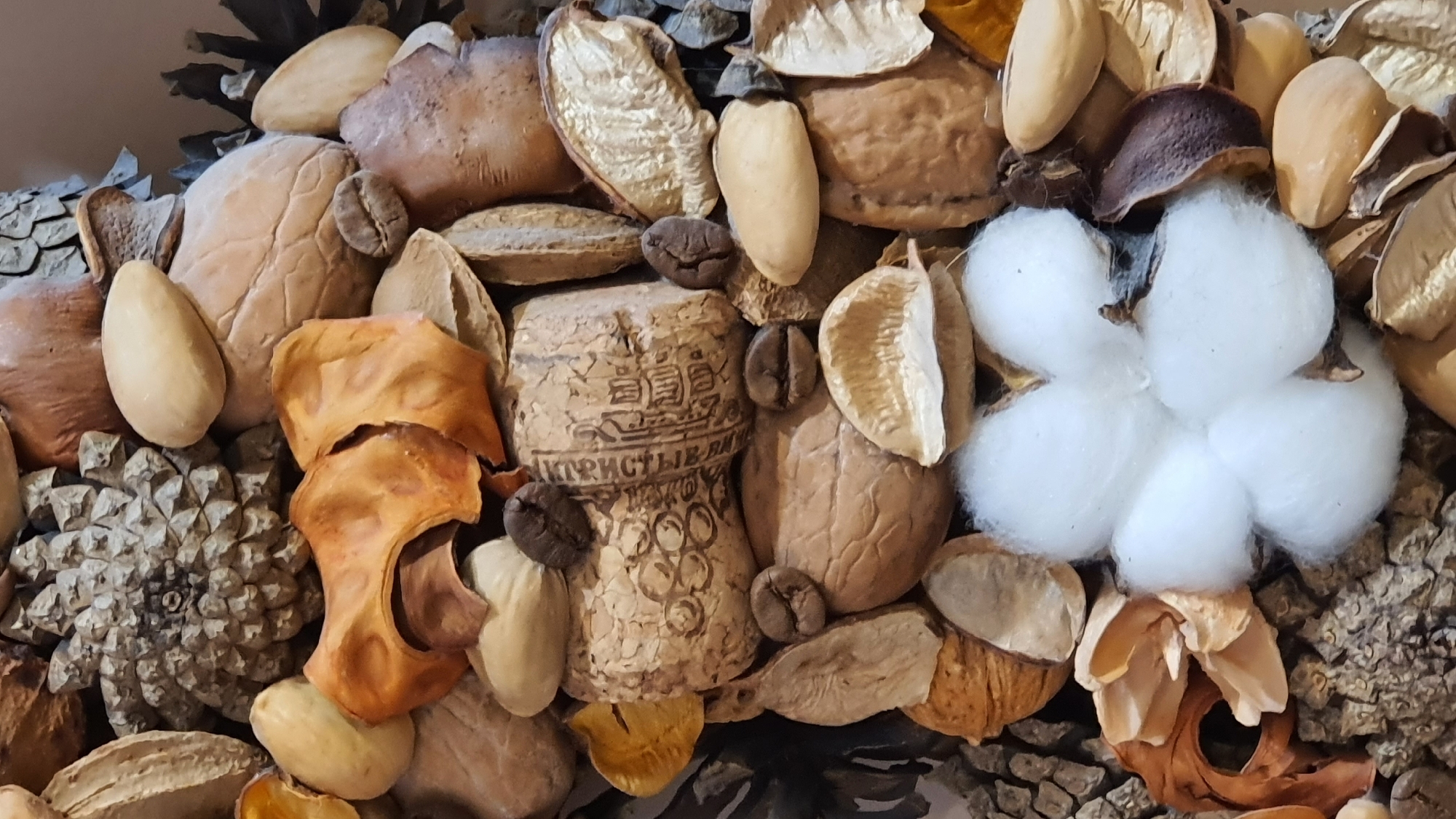 This is a close-up, top-down, vertical, still-life photograph of a decorative arrangement of natural objects. The arrangement includes a pinecone, various nuts and seeds, dried orange peel, coffee beans, and a fluffy white cotton boll. The objects are densely packed and arranged in a visually textured manner. The lighting is natural and evenly distributed, highlighting the textures and colors of the materials. The background is indistinct, suggesting a shallow depth of field. There are no people or animals present, and no specific location in Krasnoyarsk, Russia, can be identified from the image.