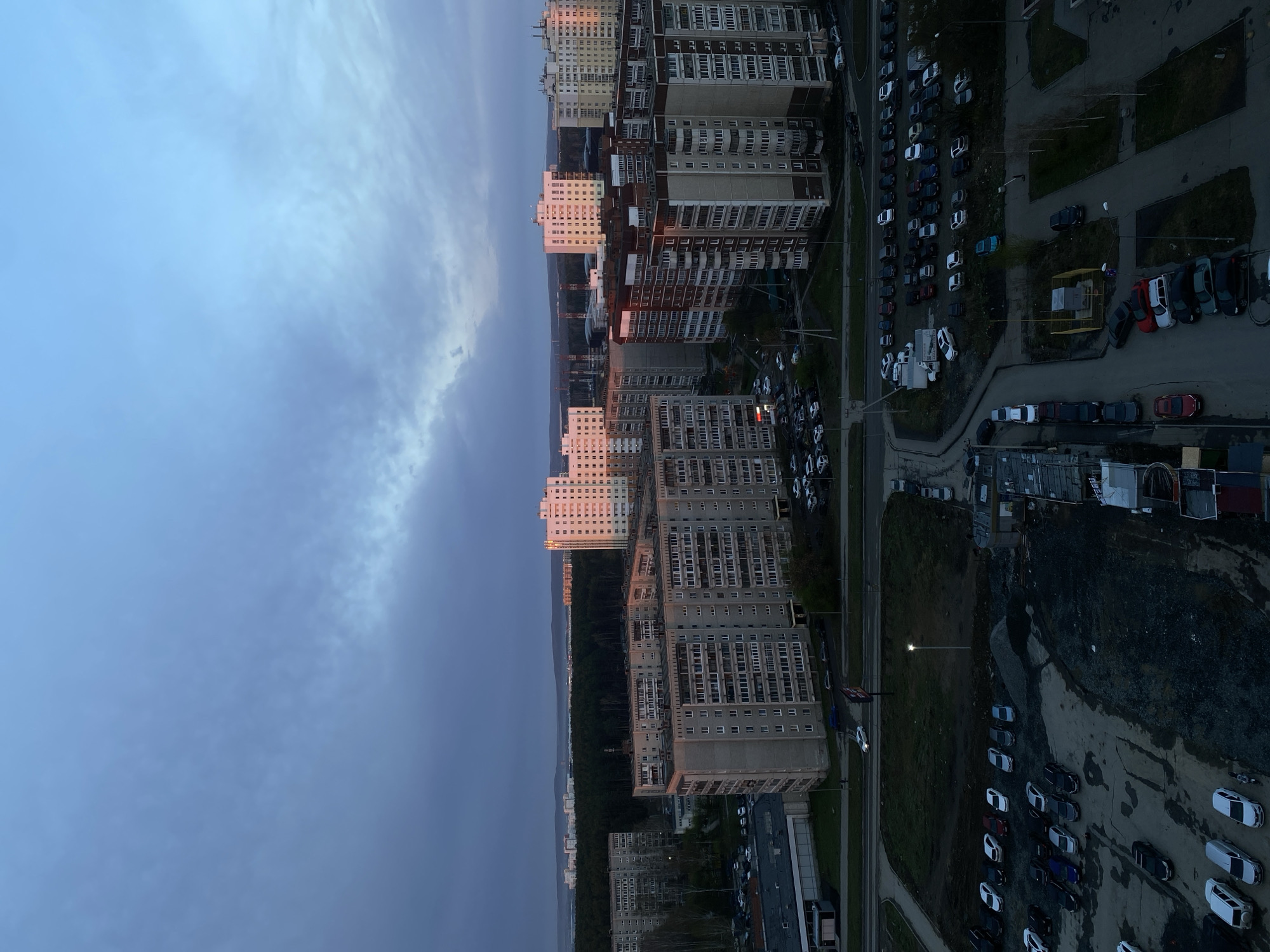 The image shows an aerial view of a residential area in Yekaterinburg, Russia. Several high-rise apartment buildings are visible, arranged in rows along a road. A large number of parked cars fill designated parking areas and along the sides of the streets. The sky is cloudy, with a transition from a lighter blue to darker, overcast tones. Between the buildings and the sky lies a strip of trees. The lighting suggests it is either dusk or dawn.