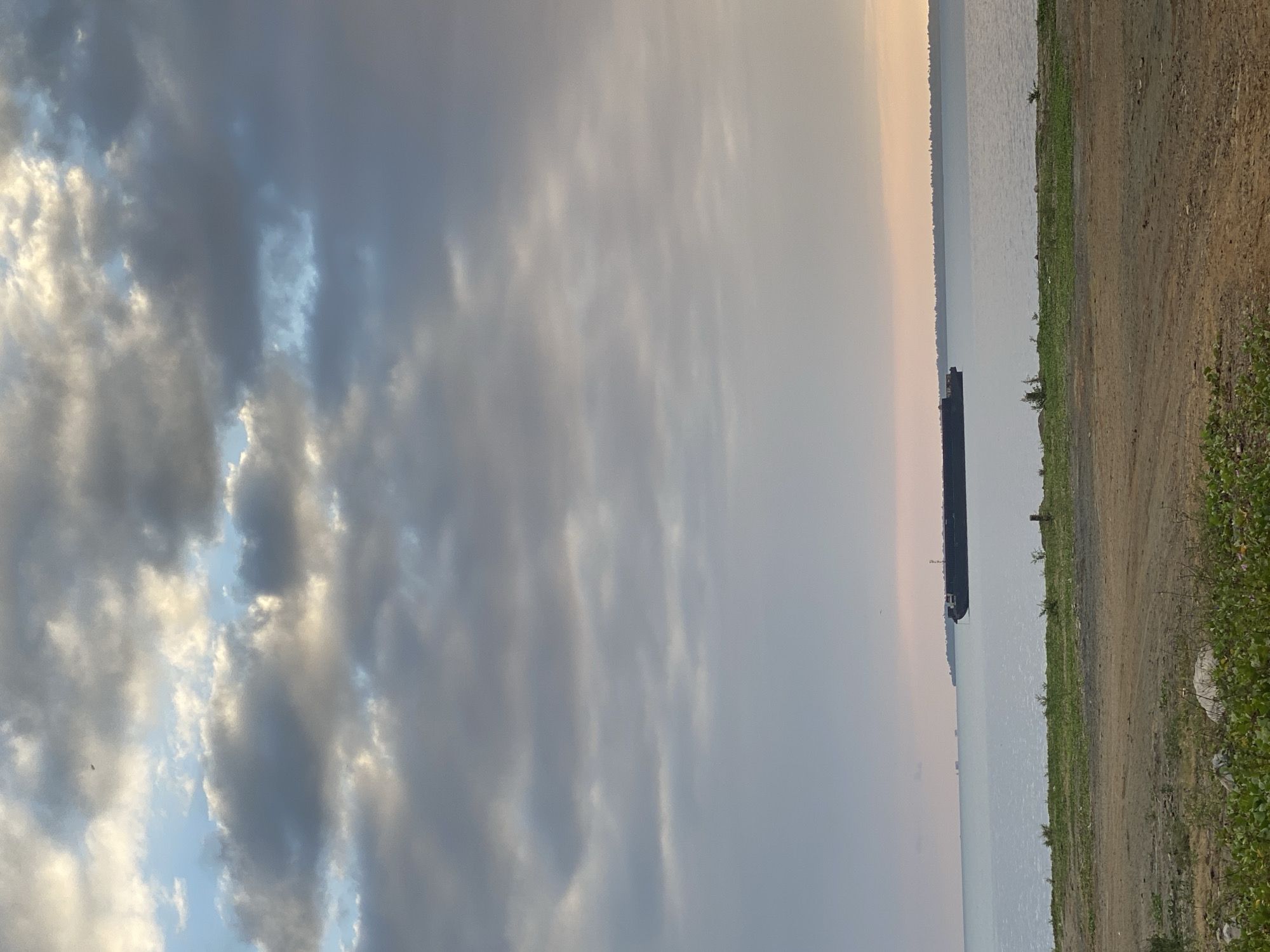 The image depicts a serene seascape with a barge on the water. The sky is filled with dramatic clouds, with patches of blue sky visible. The sea is calm, and in the distance, a faint landmass can be seen. In the foreground, there is a strip of green grass and a dirt path. The overall impression is one of peacefulness and natural beauty. The image is undated and shows no people.