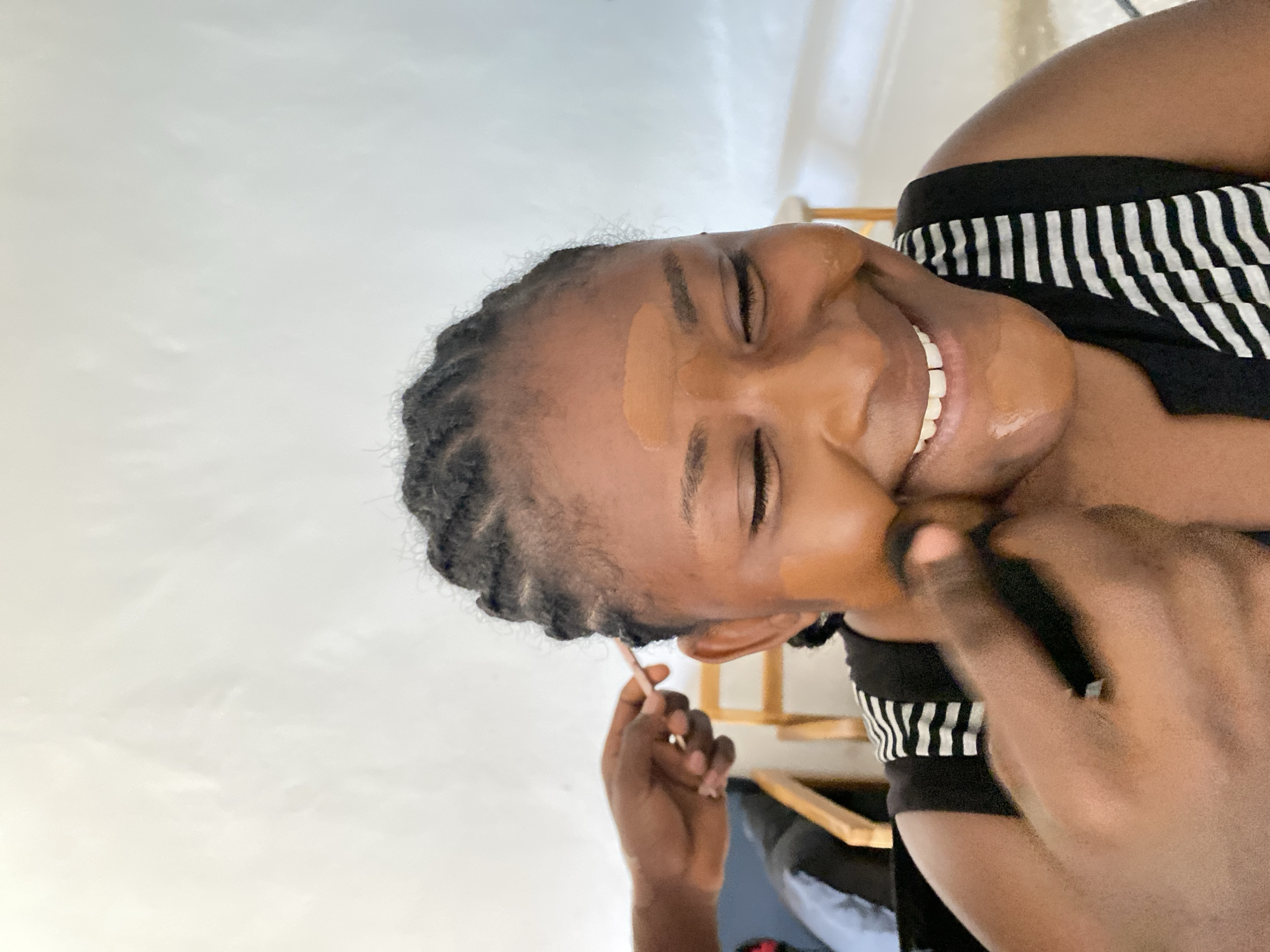 The image shows a person smiling with their eyes closed. The person has a medium skin tone and braided hair. There are patches of a lighter foundation or concealer on their forehead and cheek. Someone is applying makeup on the person, holding a brush near their hairline. The person is wearing a black and white striped top. The background is a plain white wall. Gura Top, Nigeria, is not identifiable in the image.