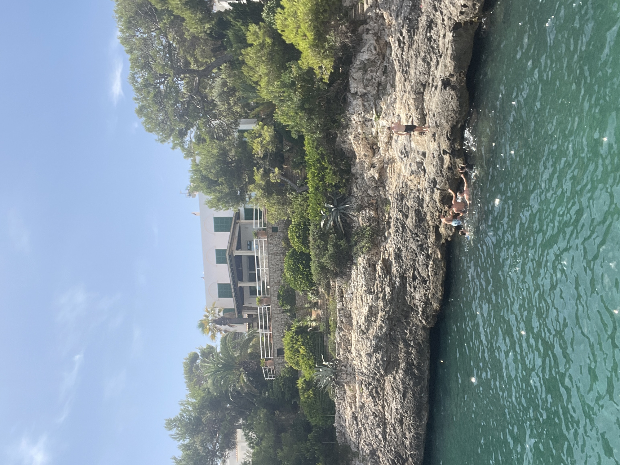 The image depicts a coastal scene in Cala D'or, Spain, featuring a body of clear, turquoise water in the foreground, with sunlight reflecting off its surface. A rocky shoreline borders the water, with several individuals engaged in swimming and interacting in the sea. One person stands on the rocks above the water, observing the scene. In the background, a multi-story white building with green shutters is visible, nestled amongst lush green trees and vegetation that covers the hillside. The building appears to be a residential property or a hotel, with balconies and outdoor seating areas. The sky is blue with a few scattered white clouds, indicating a sunny day. The overall impression is one of a serene and idyllic vacation setting.
