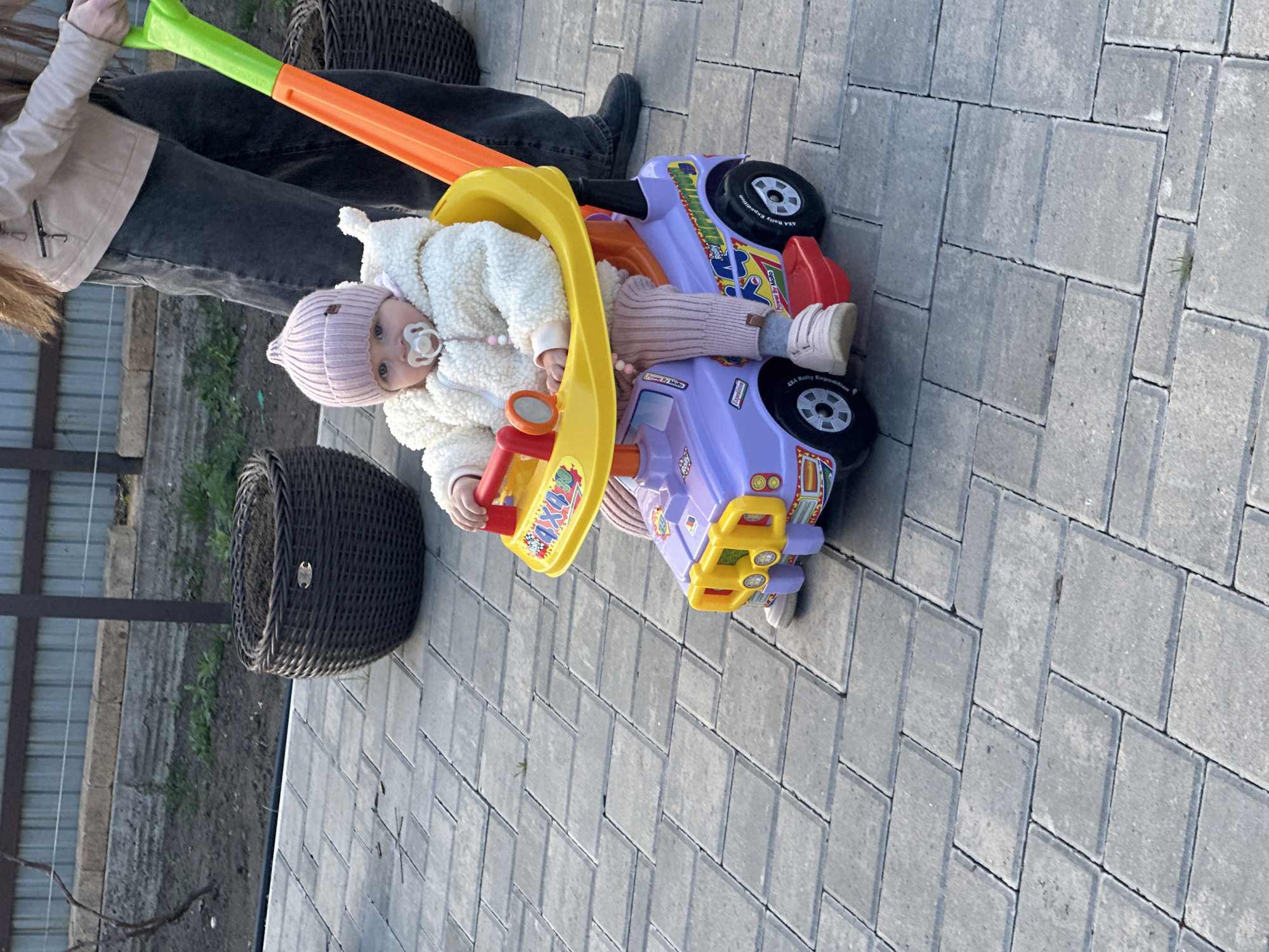 The image shows a baby in a white, fluffy jacket and a pink hat, sitting in a purple and yellow toy car with a green and orange handle. The baby has a pacifier in its mouth. An adult stands next to the car, wearing black jeans, a light-colored jacket, and black shoes. Two woven baskets are also visible, one in the background and one to the side. The scene is set on a paved area with gray brickwork. There is no evidence to suggest the image was taken in the city of Chaikovsky, Russia.