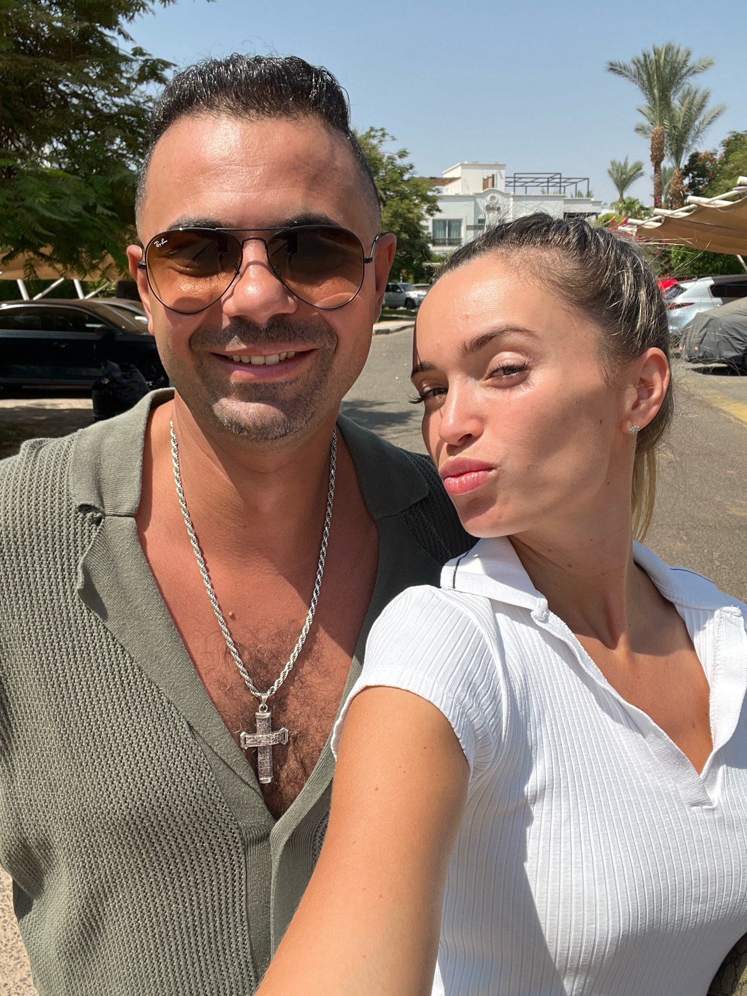 In Sharm El Sheikh, Egypt, a man and a woman are captured in a close-up selfie outdoors. The man, wearing aviator sunglasses and a green knitted shirt, has his arm around the woman. He is adorned with a prominent silver rope chain necklace featuring a cross pendant. The woman, with her hair pulled back, is looking towards the camera with her lips puckered, wearing a white ribbed polo shirt and small stud earrings. The background suggests a sunny environment with palm trees, buildings, and parked cars, hinting at a resort or residential area in the city.