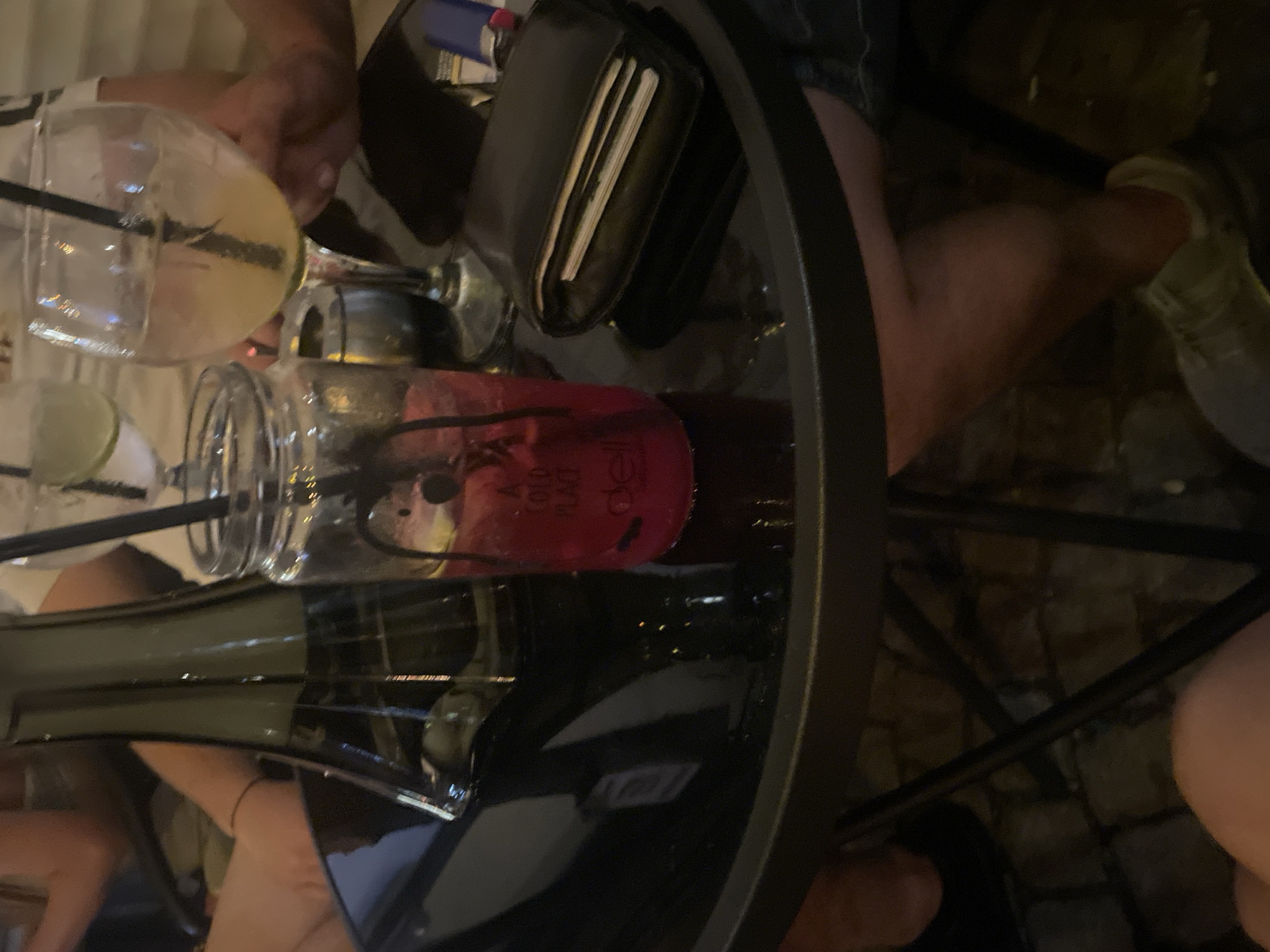 The image depicts a dimly lit outdoor or semi-outdoor social setting, likely a cafe or bar, viewed from a slightly elevated perspective focusing on a table. A dark, possibly black, round table with a glossy surface occupies the mid-ground, reflecting surrounding elements. On the table, from left to right, are: a tall, dark green glass bottle or hookah base; a mason jar-style glass containing a red beverage with ice and a black straw, bearing the text "A COLD PLACE" and "deli"; a small, dark cylindrical object resembling an ashtray; and a stemmed goblet with a yellowish beverage, ice, and a black straw. To the right of these drinks are a black leather bifold wallet with papers visible inside, and a partial view of a blue cigarette lighter.
Several individuals are partially visible. In the upper left background, a person's light-skinned arm and torso in a light-colored top are indistinctly present. In the upper right background, another person's arm is visible, seemingly holding or resting near the stemmed goblet. Below the table, a person's bare legs with light skin are visible in the lower left, while another person's leg wearing dark pants or jeans and a light-colored sneaker is visible in the lower right. The floor beneath the table consists of dark, irregular paving stones or cobblestones. The overall lighting is low, creating a relaxed ambiance. No specific interactions between individuals are discernible beyond their shared presence around the table. The location is in the city of Unknown, Unknown.