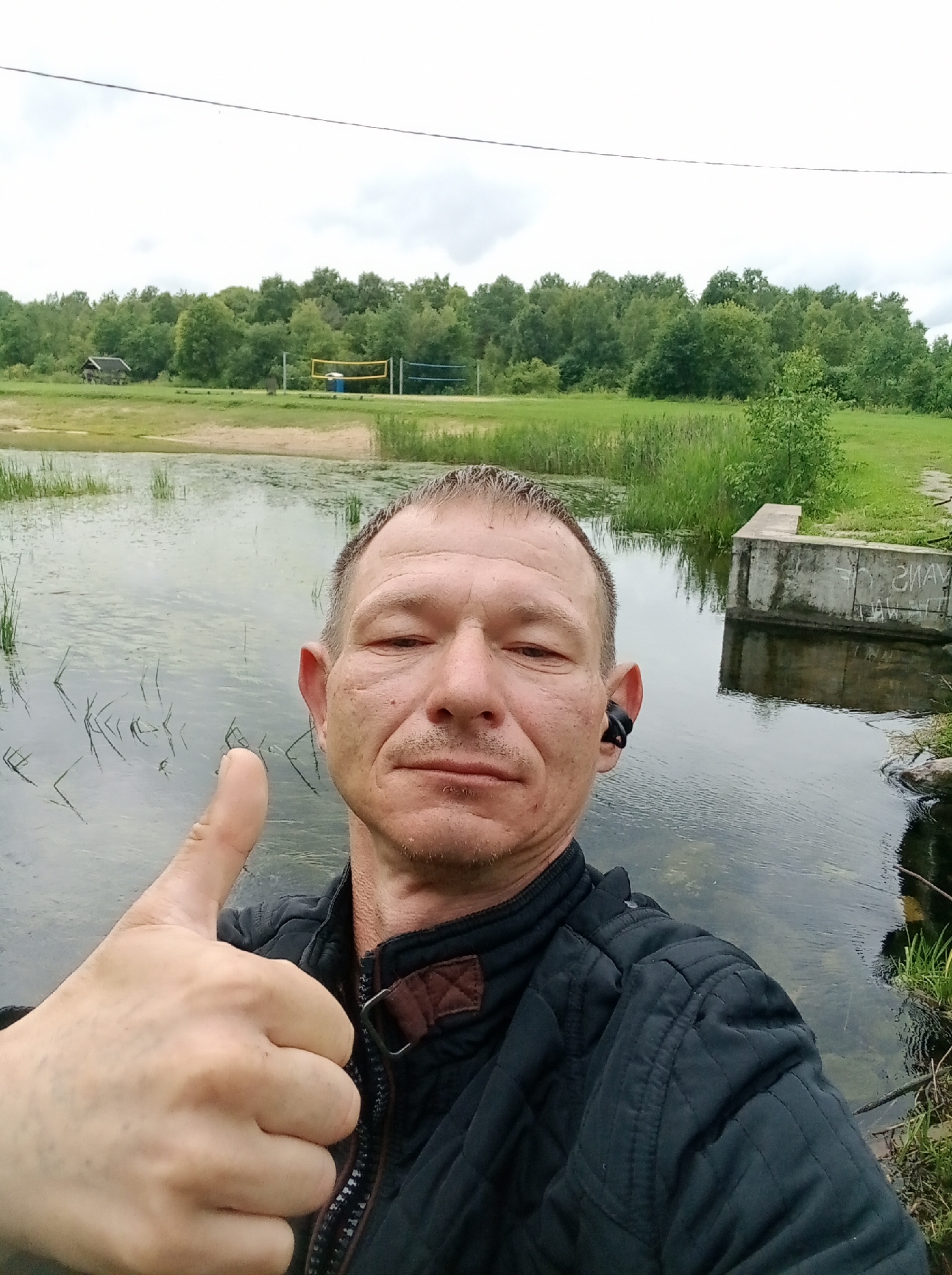 A man with short hair and a mustache takes a selfie in front of a body of water. He is wearing a dark quilted jacket and a black earbud in his right ear. He is giving a thumbs up with his left hand. In the background, there is a volleyball net with a yellow net and blue poles, and further back a dense line of trees. There is also a small wooden structure visible to the left of the volleyball net. The ground in front of the trees is grassy. The water appears to have some algae or plant growth on its surface. The sky is overcast. The location is identified as Tapa, Estonia.