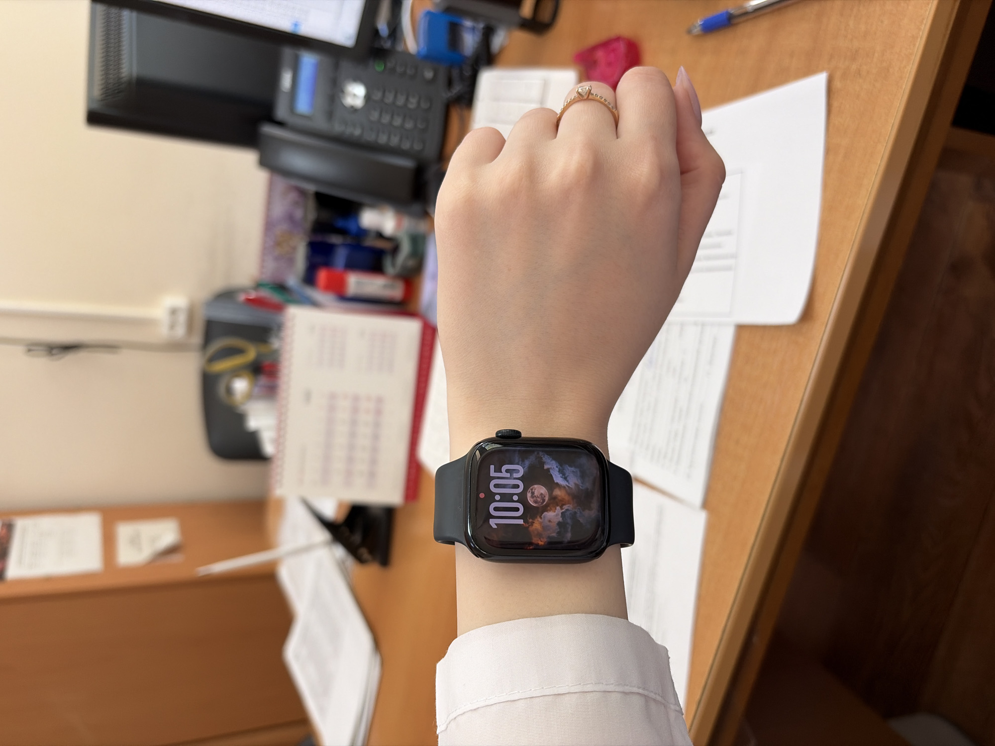A person's left wrist is displayed wearing a black smartwatch with a screen showing a digital clock reading 10:05 and a background image with a moon. The person is also wearing a gold ring on their ring finger. The wrist is placed on a desk covered in paperwork, with office equipment such as a desk phone, a calendar, and stationery in the background. The location is likely an office setting in Chaikovsky, Russia, based on the source data.