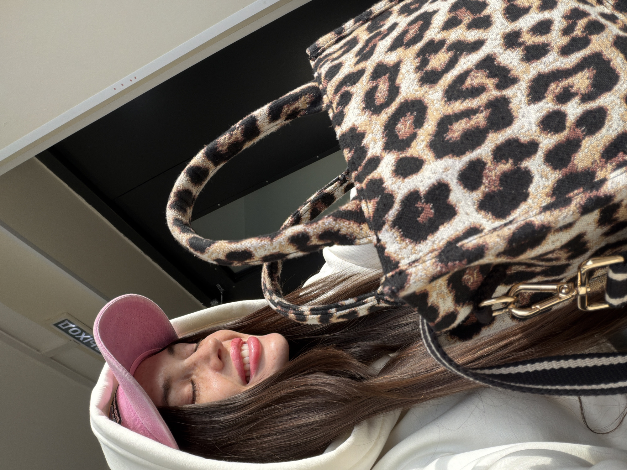 A young woman, angled sideways, smiles with her eyes closed. She is wearing a light pink baseball cap and a white hoodie. Above her is a large leopard print bag with gold hardware and a black and white striped strap. The background suggests an interior space with a window. The sign on the upper left says "EXIT" in reverse, in English. The location is unlikely to be Saint Petersburg, Russia, since signage in Russia would be in Russian.