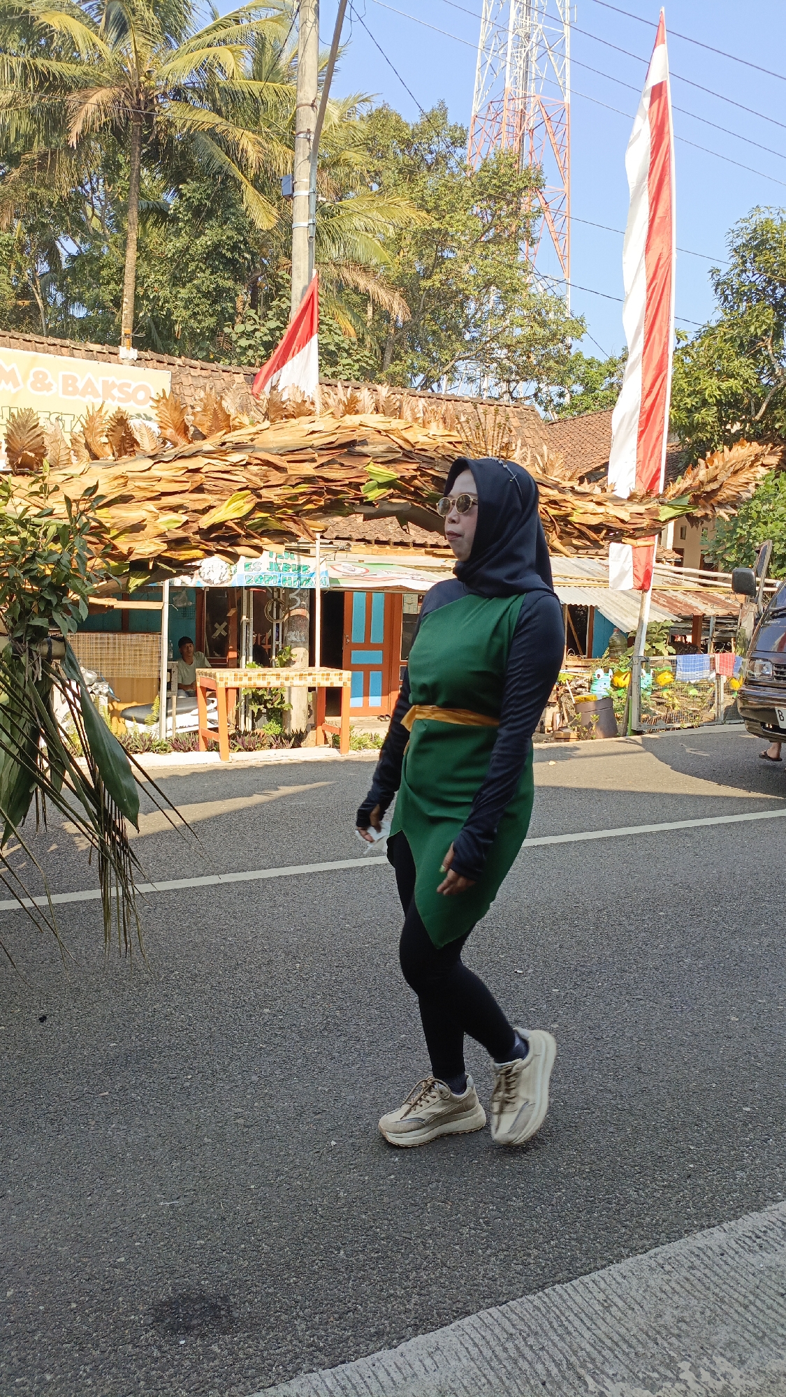 The image depicts a woman in a green tunic and black leggings, wearing a black hijab and sunglasses, walking on a paved road. She is also wearing light-colored athletic shoes. She is seen from the side, facing left, and appears to be in motion. Behind her, there is a building with a brown tiled roof adorned with what appear to be dried leaves or palm fronds. Indonesian flags, red and white, are visible on poles in front of the building. A telecommunications tower stands tall in the background, amidst lush green trees and palm trees. The sky is clear and blue, indicating a sunny day. The context provided indicates the location as Krajan Lor, Indonesia. The scene appears to be a street in a rural or semi-rural area, possibly a marketplace or a roadside establishment, given the building and the flags. There is a suggestion of commercial activity, with a sign on the building that reads "M & BAKSO." A person is seated inside the building, and a vehicle is parked on the right side of the road.