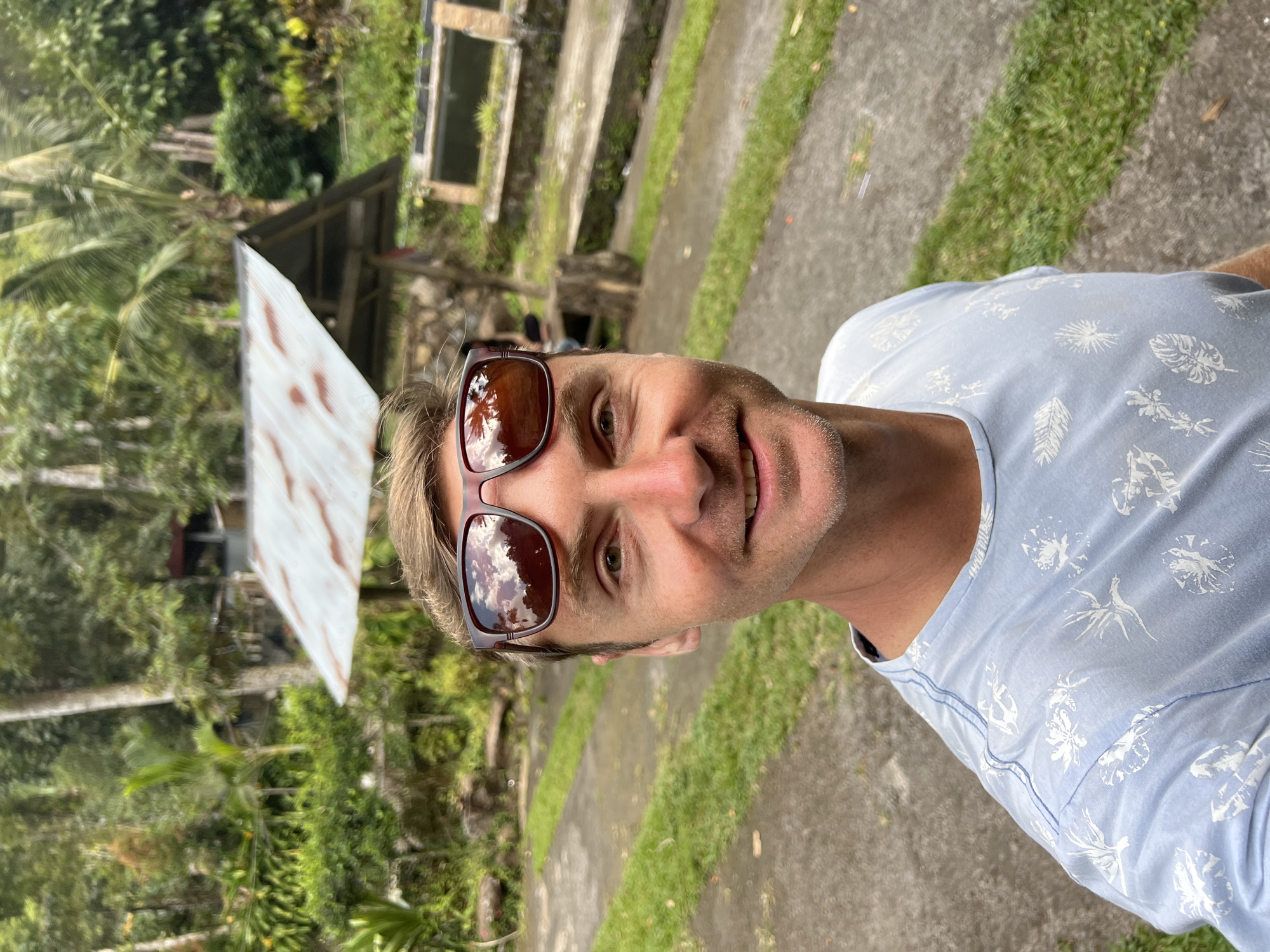 A man with blonde hair and a light blue t-shirt with a floral print is taking a selfie outdoors. He is wearing sunglasses on his head. The background shows a lush, green environment with trees and foliage, as well as some structures that appear to be part of a park or resort. There are concrete pathways and grassy areas, and a small, rustic-looking shelter with a corrugated metal roof. The overall impression is a casual, outdoor setting in a tropical or subtropical climate. The presence of what appear to be fish ponds in the background suggests the location may be a tourist attraction or a place focused on aquaculture. The man's expression is relaxed and smiling, indicating he is enjoying himself. The photograph seems to capture a moment of leisure and sightseeing.