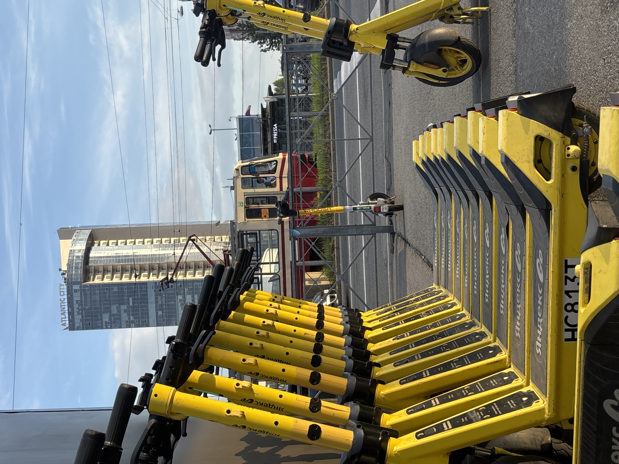 The image shows a line of Yandex Go electric scooters parked on a street in Saint Petersburg, Russia. A tram is visible in the background, along with a building labeled "ATLANTIS CITY." A scooter labeled "WHOOSH" is also present. The sky is partly cloudy.