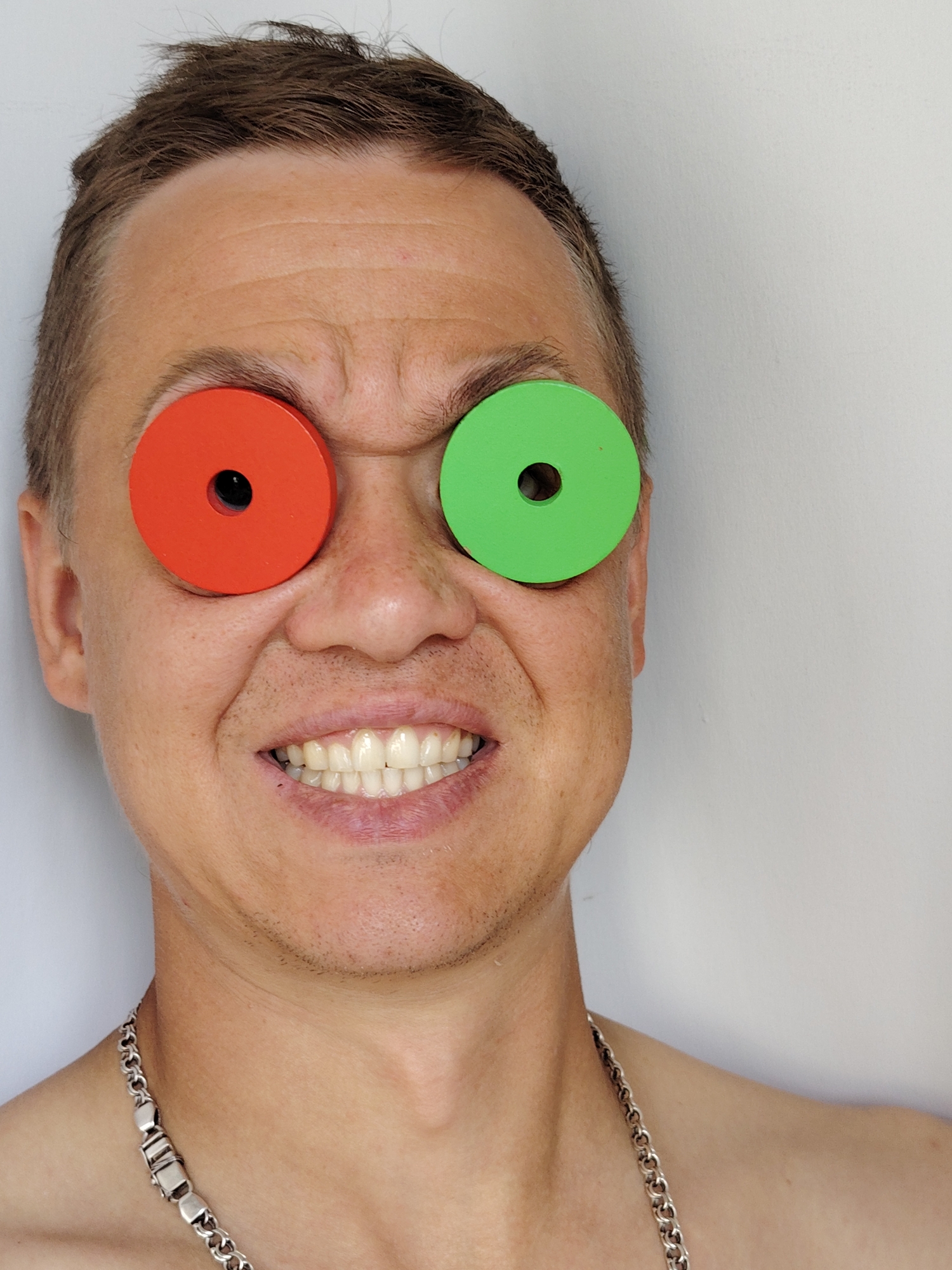 A man's face is positioned in the center of the frame, facing forward. He has short, brown hair and appears to be smiling with his teeth showing. Red and green circular objects, each with a hole in the center, are placed over his eyes, obscuring them from view. He is wearing a thick, silver chain necklace. The background is a plain, light-colored wall. There is no discernible location within the city of Chaikovsky, Russia.