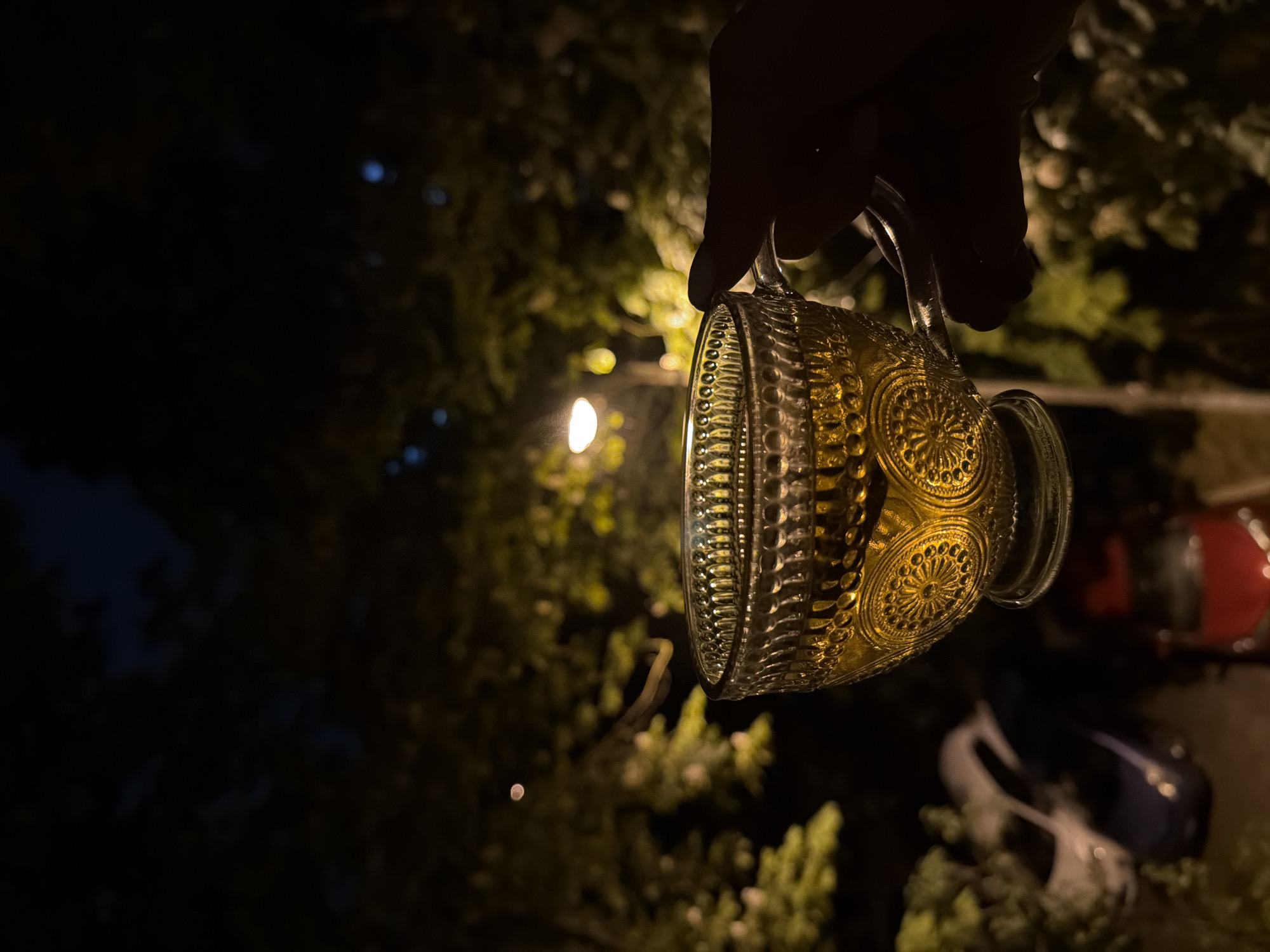 A hand holds a glass mug with a decorative pattern containing a yellow liquid. The background is dark with blurred shapes of trees and two cars. A single light is visible. The location is Unknown, Unknown.