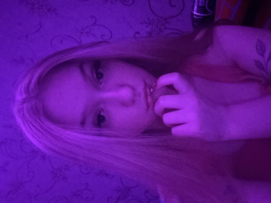 The scene depicts an interior space, reportedly located in the city of Saratov, Russia. A single female individual is positioned centrally, facing directly towards the camera under predominant intense purple lighting. Her long, light-colored hair, appearing pinkish-purple under the illumination, falls over her shoulders. She exhibits a neutral expression, with her eyes open and looking forward, and her lips slightly parted and glossy. Her right hand is raised, with fingers curled, and the index finger touches her lower lip; her fingernails are painted red. She wears a top that appears reddish-pink under the purple light, possibly a tank top or camisole. A tattoo of a leafy branch design is visible on her left forearm/upper arm. The background consists of a wall covered in patterned wallpaper, featuring dark, ornate floral or scroll motifs on a lighter, purple-tinted base. A dark, partially visible object consistent with furniture or a bedside item is present in the lower right corner. The environment suggests a residential or private room.