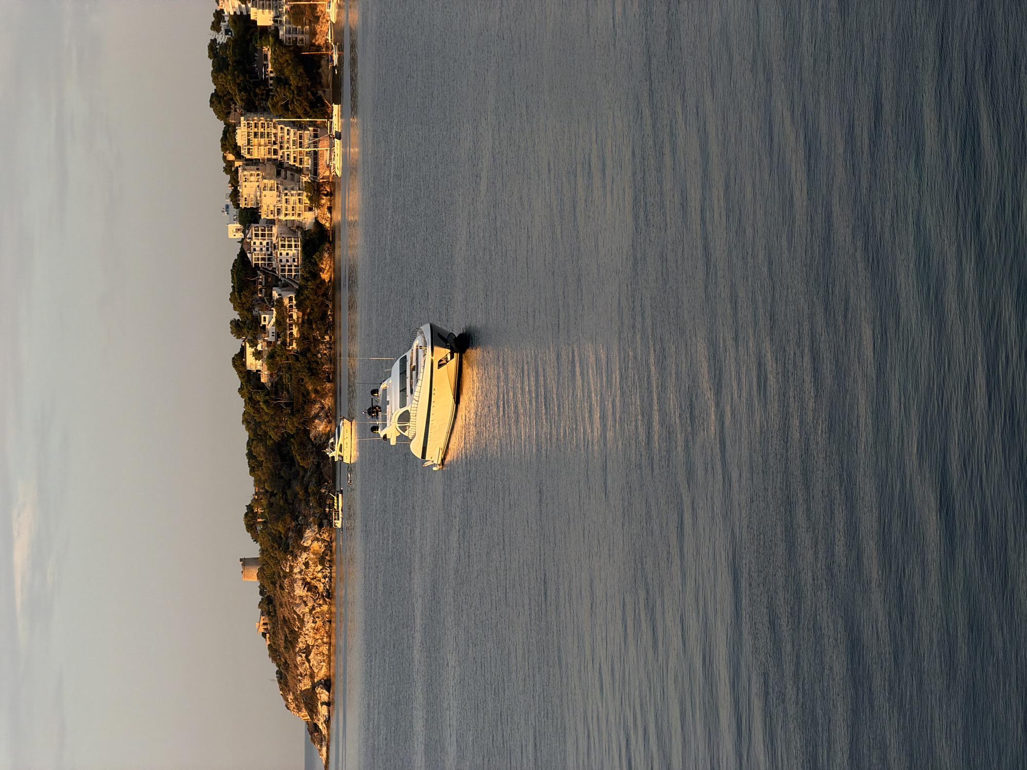The image depicts a serene seascape in Palmanova, Spain, bathed in the warm, golden light of what appears to be late afternoon or early morning. The foreground is dominated by the calm, rippling water of the sea, reflecting the sky and surrounding landscape. In the mid-ground, a sleek, white motor yacht is anchored, its hull gleaming in the sunlight.
In the background, the coastline of Palmanova is visible, characterized by a lush green hillside dotted with trees. Perched atop the hill is an old, circular stone tower, likely a historical landmark. Nestled along the slope are numerous white buildings, appearing to be apartment complexes or hotels, with multiple balconies and windows. The architecture suggests a Mediterranean coastal town. Several other smaller boats and yachts are also visible anchored in the bay, adding to the sense of leisure and activity. The sky above is a pale, hazy blue with subtle clouds, indicating fair weather. The overall impression is one of tranquility and picturesque coastal scenery.