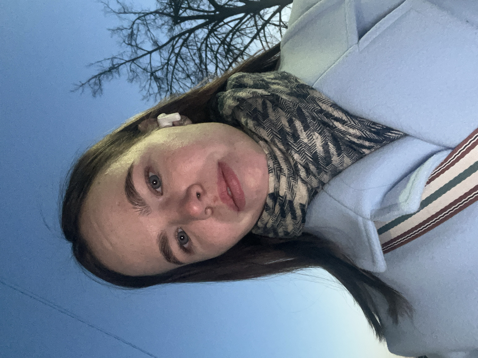 A young adult female is depicted in a selfie-style portrait, looking directly at the viewer. She has long, dark brown hair, light-colored eyes, and a neutral facial expression. A white wireless earbud is visible in her left ear. She is wearing a light blue coat with a visible collar and a patterned scarf around her neck, featuring a chevron or herringbone design in dark and light tones. A bag strap with vertical stripes in brown, white, and green is visible across her left shoulder. The background consists of a clear, dark blue sky, suggesting dusk or dawn conditions. Bare tree branches, silhouetted against the sky, are visible on the right side of the frame. A faint, thin line, possibly a wire or distant structure, is discernible in the upper left portion of the sky. The specific location in the city of Chaikovsky, Russia, cannot be determined from the visual information provided in the image.