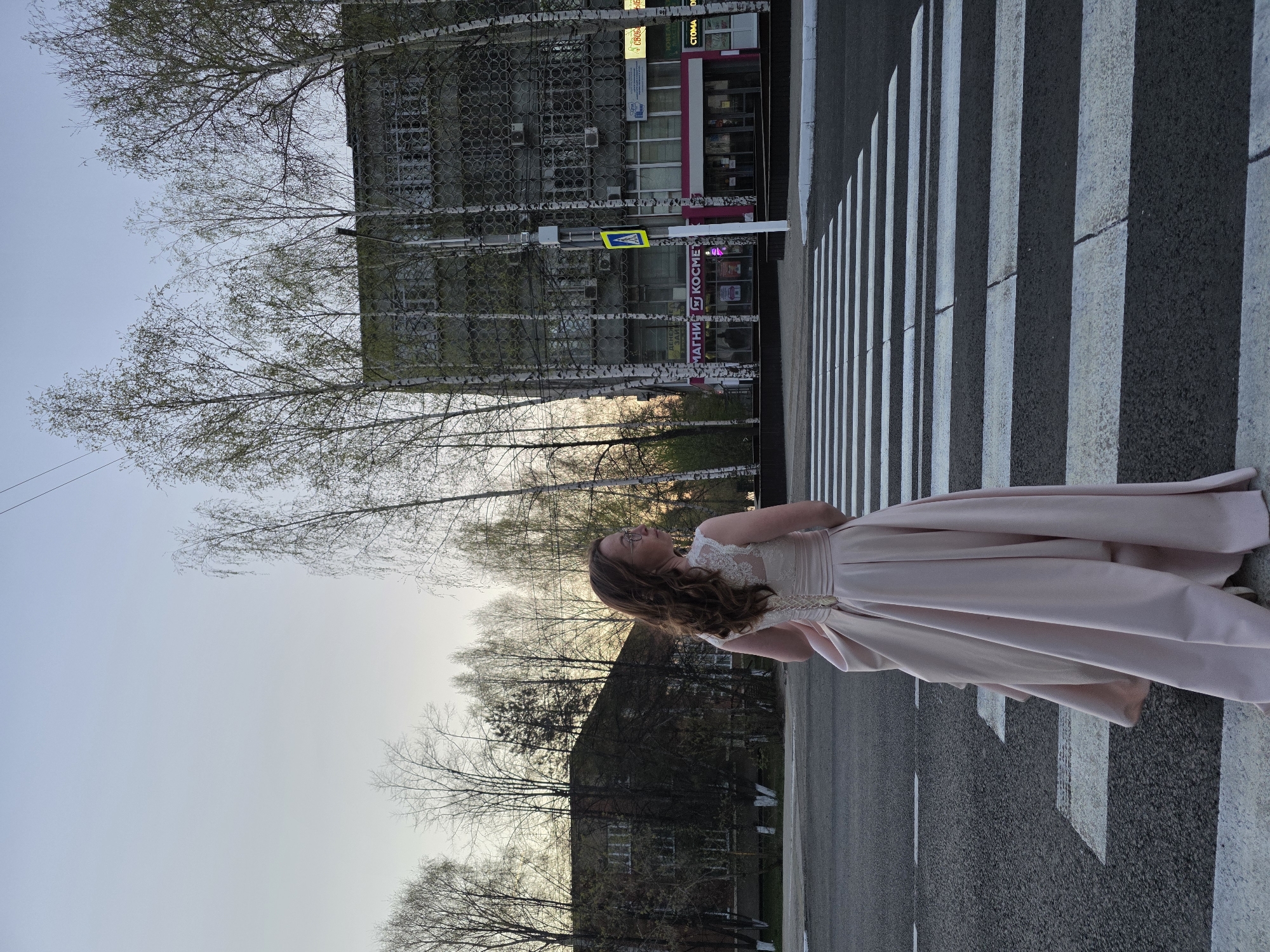 A woman in a light pink dress stands on a zebra crosswalk in the city of Chaikovsky, Russia. She has long, wavy brown hair and is looking up at the sky. Her arms are crossed. Behind her is a grey multi-story building and bare trees. There is a sign with the word "КОСМЕТИКА" visible on one of the buildings. The sky is light blue with hints of pink.