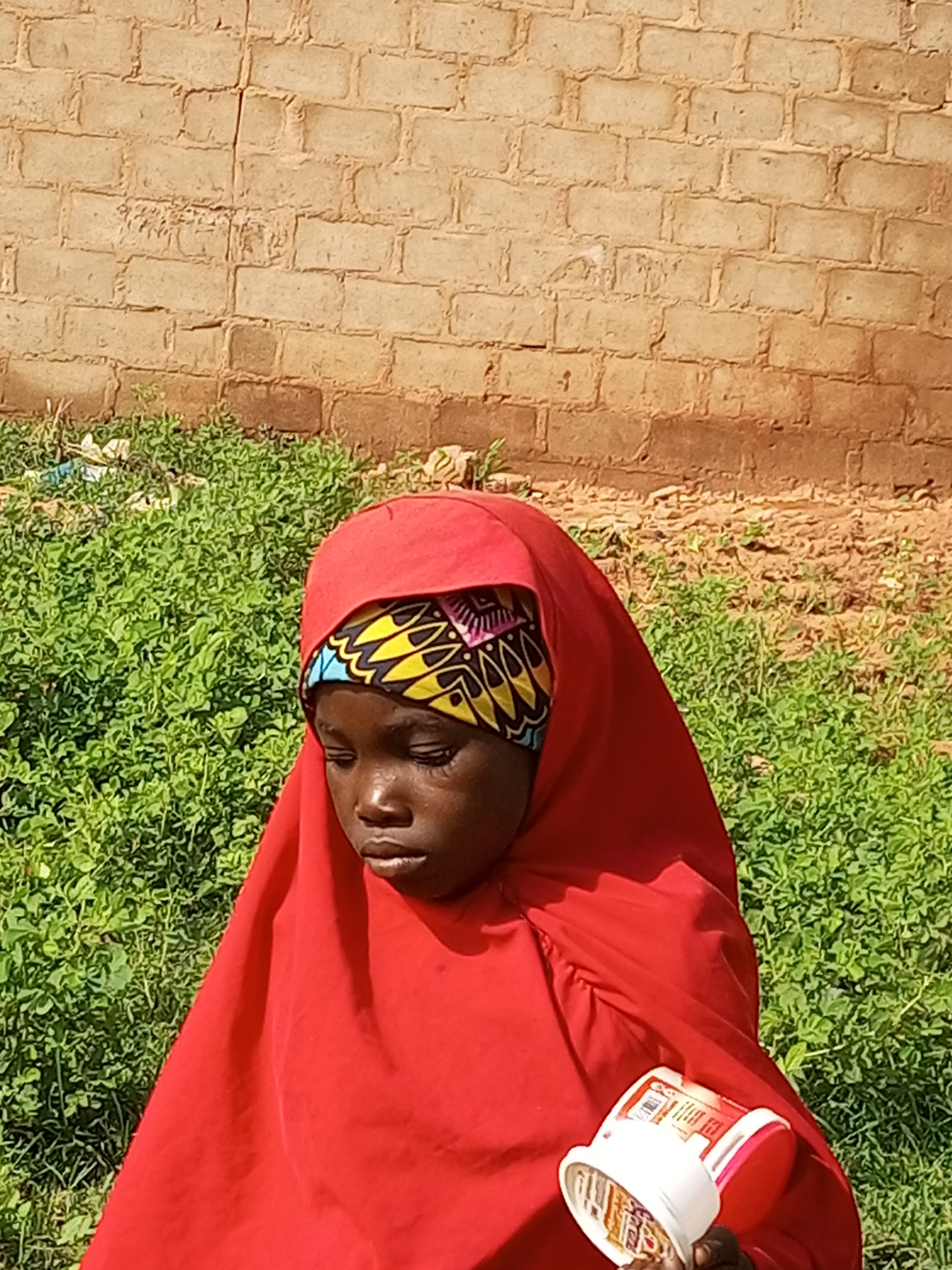 A young Black girl wearing a red hijab with a colorful headscarf underneath looks down to her right while holding a small white and red plastic container in her right hand. She is positioned in front of a brick wall and a patch of green vegetation. The background consists of a light brown brick wall with visible mortar and a grassy area with some litter. The lighting suggests it is daytime. The girl's expression is somber.