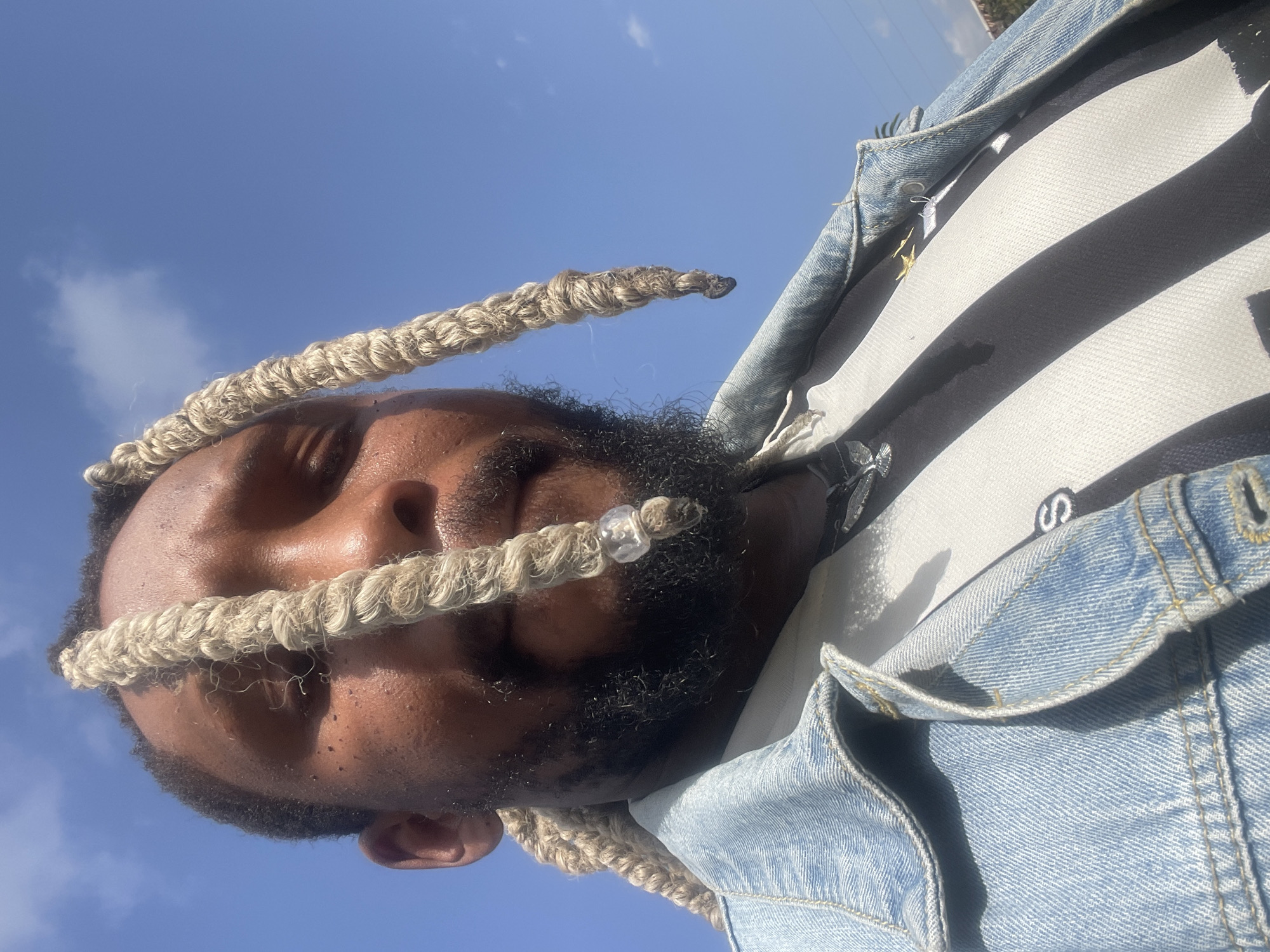 This media file features a close-up, low-angle shot of a Black man outdoors against a bright blue sky with scattered clouds. The man has his eyes closed, appearing to be relaxed or basking in the sun. He has a beard and light-colored braided hair, with one braid adorned with a clear bead. He is wearing a denim jacket over a striped shirt. The lighting suggests it is daytime, likely sunny. The scene is set in Jos, Nigeria.