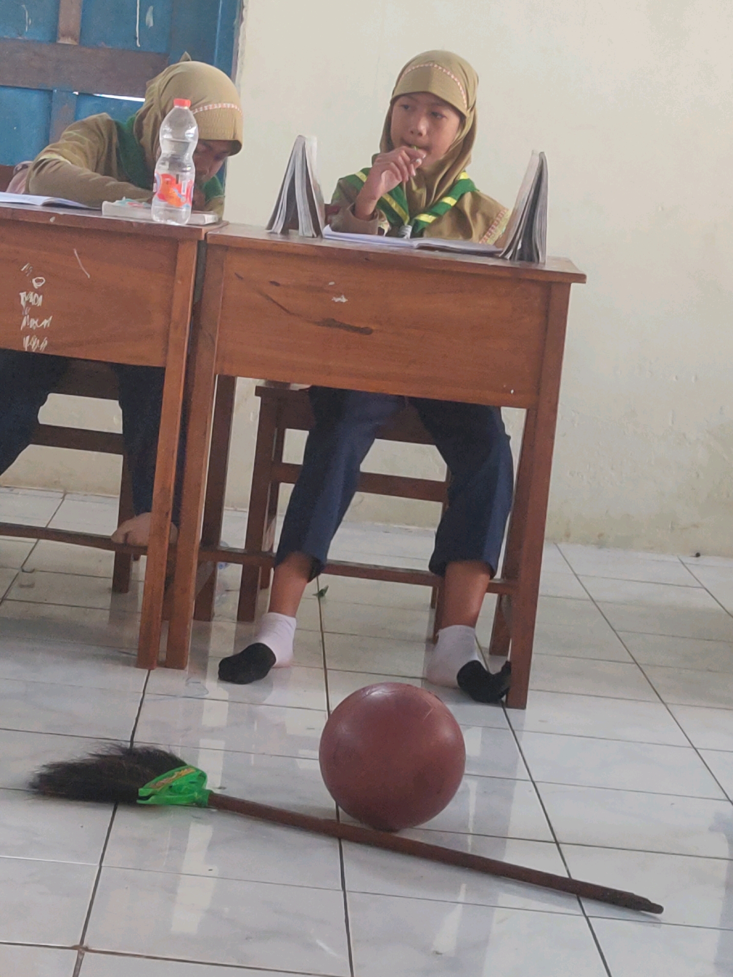 In a classroom setting in Payaman, Indonesia, two young girls are seated at wooden desks. The girl on the left, wearing a tan hijab and a brown and green uniform, is looking down at her books, with a bottle of water on her desk. The girl on the right, also in a tan hijab and a similar uniform, is looking towards the viewer and has her hand near her mouth. Both girls are wearing dark trousers and white socks. In the foreground, a red ball and a broom are lying on the tiled floor. The classroom walls appear to be painted white.