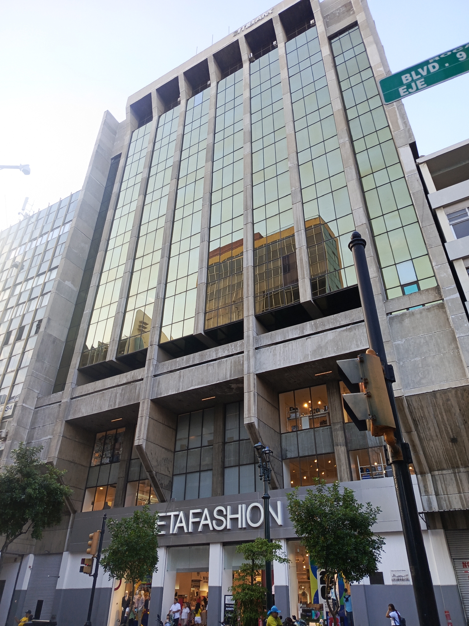 This is a ground-level, eye-level, slightly angled shot of a modern building in Guayaquil, Ecuador. The building is tall and features a facade of reflective, golden-tinted glass panels divided by concrete beams. The lower floors house a retail store called "TETA FASHION," indicated by large white lettering. Several people are visible near the store entrance, some standing and some seated. A traffic light pole stands prominently in the foreground to the right of the frame, with a green street sign that reads "BLVD. 9 DE OCTUBRE EJE". Trees and street lamps are also present. The sky is bright and clear, suggesting daytime. The architecture is Brutalist-inspired, with strong geometric lines and exposed concrete. Reflections of other buildings are visible in the glass facade.