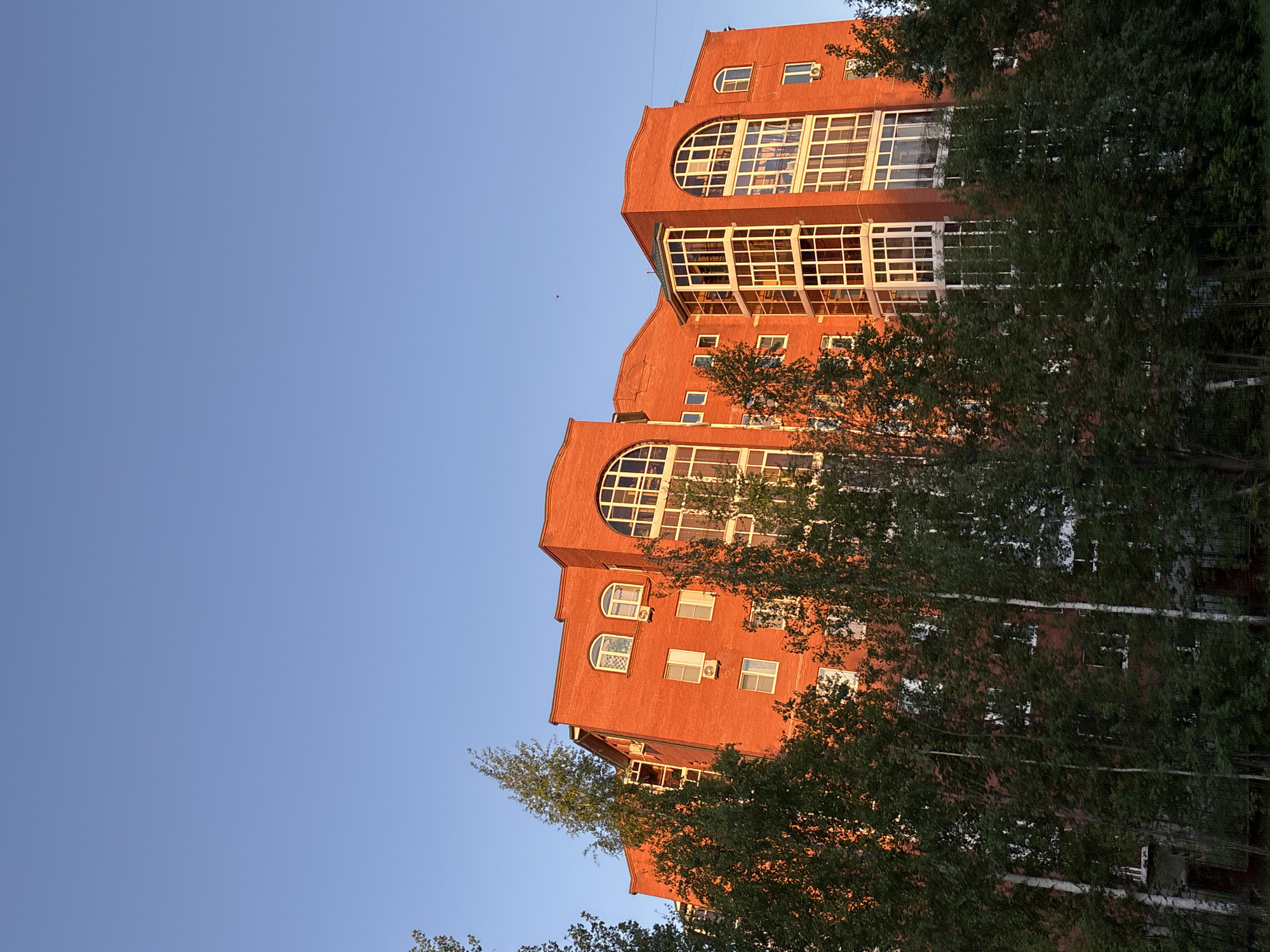 The image captures a multi-story brick building in what appears to be a residential area in Chaikovsky, Russia. The building is reddish-orange in color and features several arched windows and balconies. Trees partially obscure the view of the building. The sky is clear and blue. The photograph is taken from a low angle.