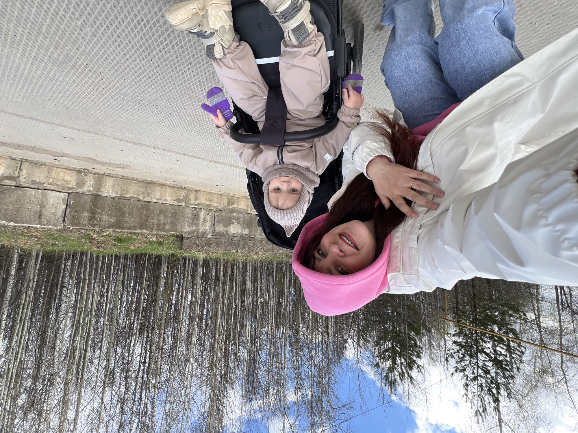 A photograph taken in Noviy Urengoy, Russia, shows a woman and a baby in a stroller, presumably a selfie. The woman wears a pink hooded sweatshirt, a white jacket, and jeans. She's holding her hand up, possibly taking the photo. The baby is in the stroller, wearing a beige jumpsuit, matching hat, and purple mittens. The background shows a concrete walkway or bridge and a line of bare trees. The sky is visible with blue patches and clouds through the trees. The image is rotated, making the ground appear to be above them.