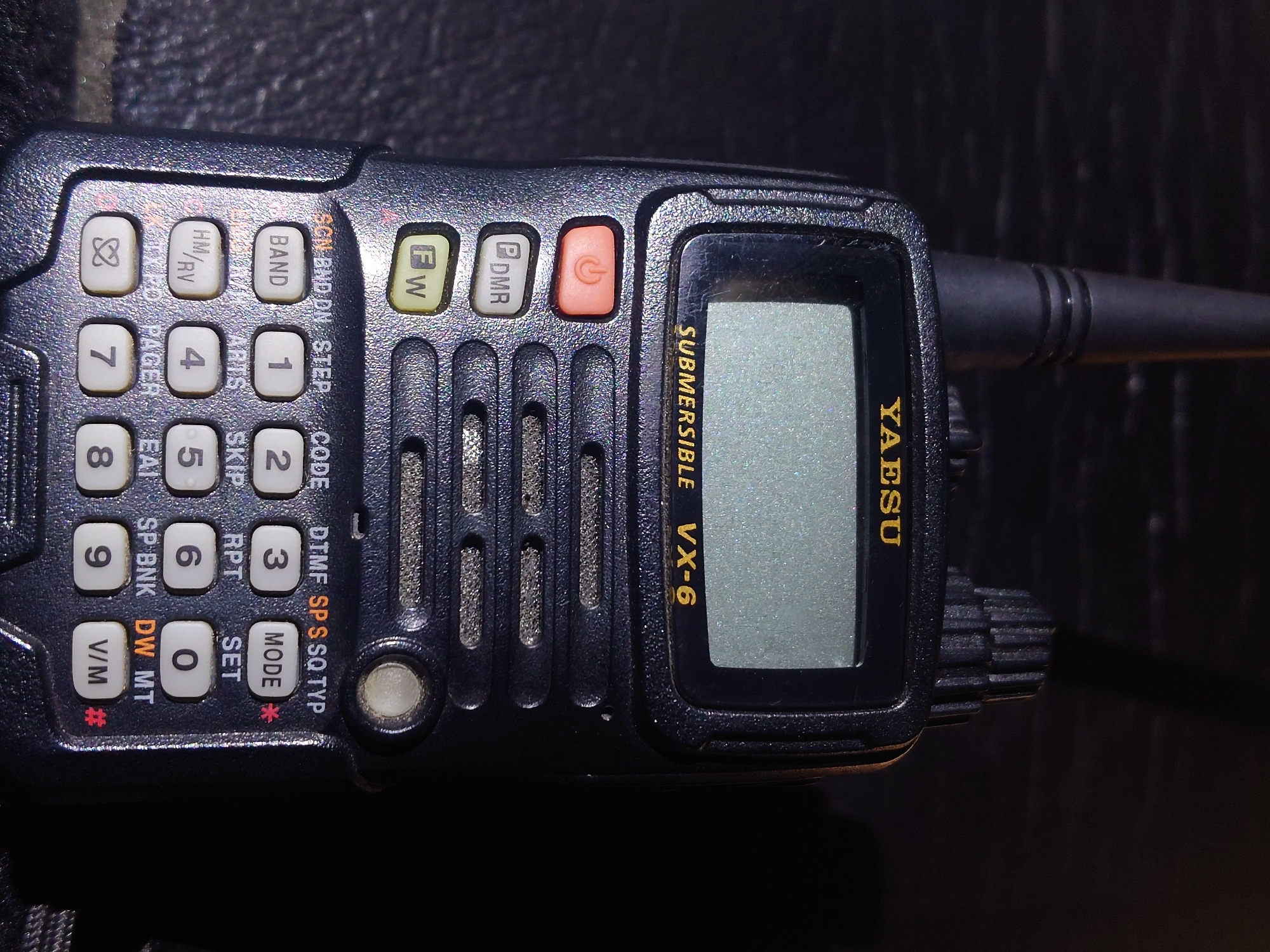 The image shows a close-up, low-angle, indoor shot of a black Yaesu VX-6 submersible handheld radio. The radio is oriented vertically and is placed on a dark, textured surface, possibly a table or fabric. The lighting is dim, with a spotlight effect highlighting the radio. The radio has a prominent antenna at the top, a grayscale LCD screen displaying the text "SUBMERSIBLE VX-6" and "YAESU" in yellow lettering. Below the screen are several buttons, including a red power button with a power symbol, a button labeled "DMR", and another labeled "FW". A numeric keypad with functions like "BAND", "STEP", "CODE", "DTMF", "MODE", "SET", "HM/RV", "ARTS", "SKIP", "RPT", "SP BNK", "DW", "MT", "PAGER", "EAI", "V/M" and numbers 0-9 are visible. A circular, translucent button with a white light is located to the right of the keypad. There are no people or discernible actions occurring in the image. The environment is dark and indistinct, suggesting an indoor setting with minimal ambient light. There is no information in the image to indicate a location in Sokolohirsk, Ukraine.