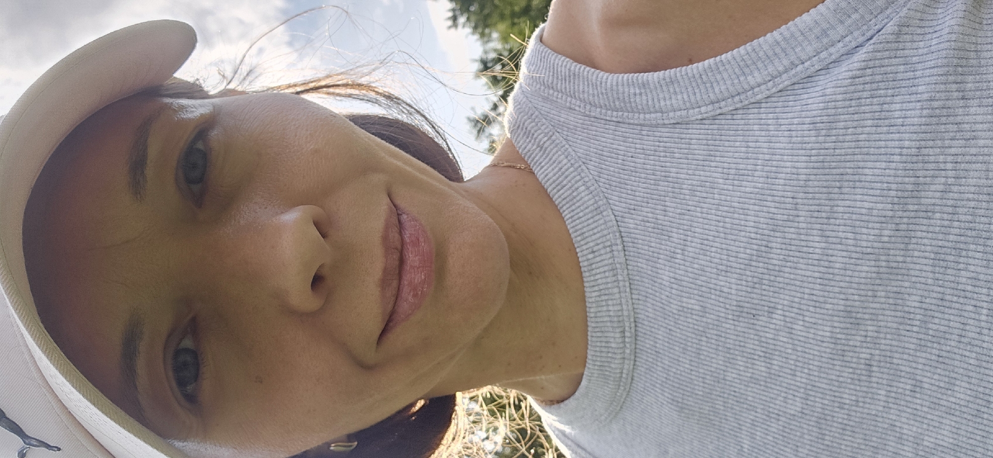 The image shows a close-up, eye-level, outdoor, frontal portrait of a woman wearing a light-colored baseball cap and a light gray ribbed tank top. She has blue eyes, fair skin, and her lips are slightly parted, revealing a subtle smile. The background features trees and a cloudy sky. The lighting suggests it is daytime. The image does not contain any information about the location in Мазалаўскі сельскі Савет, Belarus.