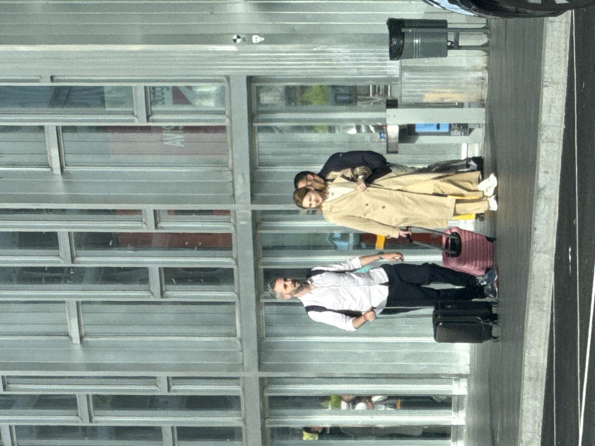 The image depicts three individuals on an outdoor paved walkway in front of a building with a glass facade. A man with a beard, wearing a white collared shirt and dark trousers, is pulling a black wheeled suitcase. To his right, a woman in a light-colored trench coat and matching trousers is walking, also with a pink wheeled suitcase. Behind her, another person, partially obscured, is walking. The building's facade is composed of vertical metal panels and numerous rectangular glass panes, some of which reflect the surrounding environment. In the background, signs are visible through the glass, including one with "AVIS" and "Budget" text. A black trash receptacle is situated to the right of the individuals. The scene appears to be an airport terminal or a similar transportation hub, potentially at Josep Tarradellas Barcelona–El Prat Airport (BCN) in El Prat de Llobregat, Spain, given the context and common architectural styles for such facilities. The lighting suggests it is daytime.