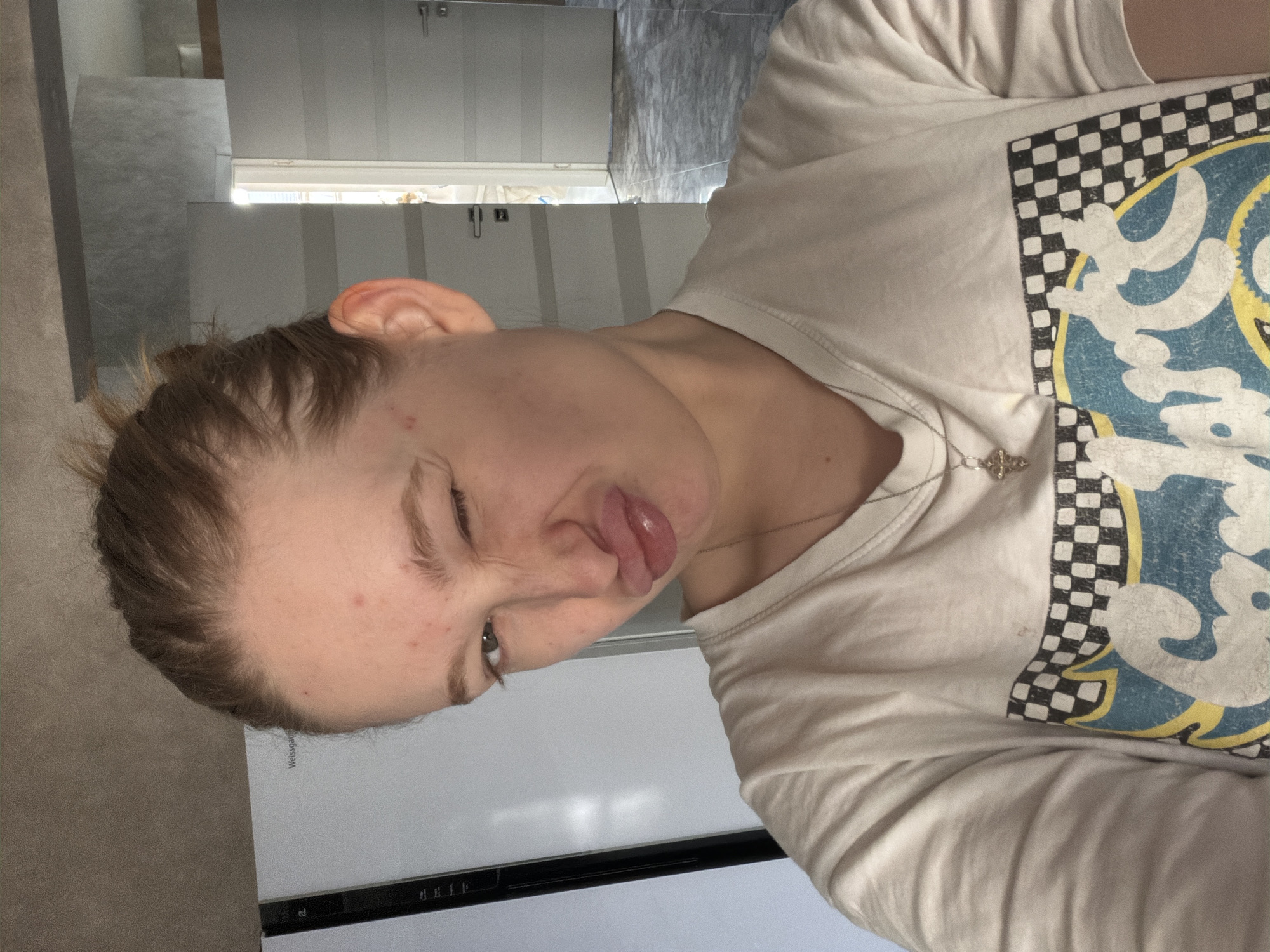 A Caucasian female with light brown hair and skin, wearing a white t-shirt with a checkered and blue graphic print and a silver necklace with a cross pendant, is making a silly face, sticking out her tongue and winking. She is indoors, likely in a residential setting, given the glimpse of a kitchen countertop and cabinets in the background. The location is Rossiyskiy, Russia.