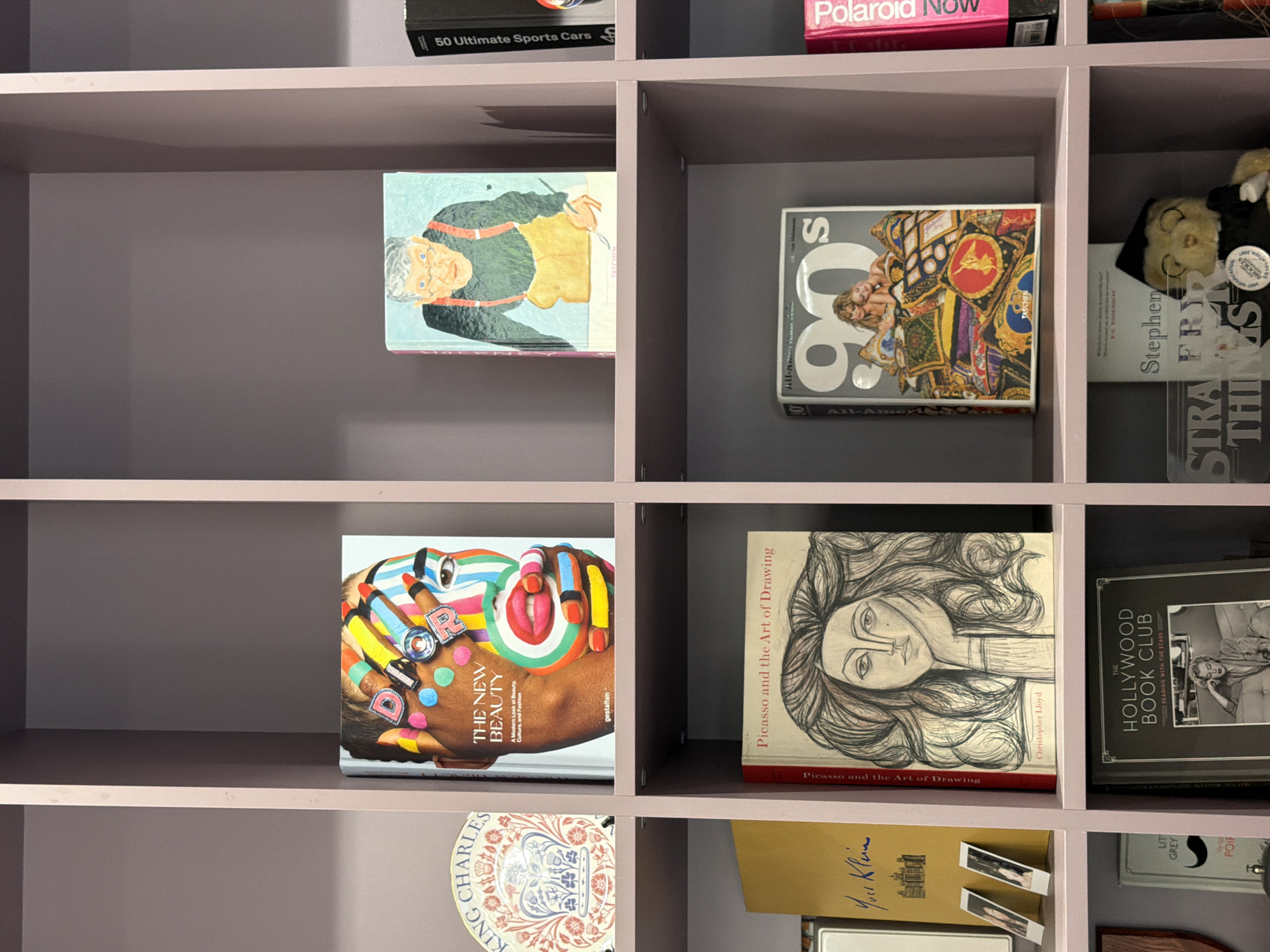 The image shows a section of a pink bookshelf with several books and decorative items arranged on its shelves. The books are of various sizes and genres, including art books and fashion magazines.
On the top shelf, to the left, is a book with a colorful abstract design on its cover, featuring close-ups of eyes and lips. To its right is another book with a painting of an older man with glasses and suspenders. Further to the right, a black book with white text is partially visible.
The middle shelf contains a yellow book on the left, followed by a book titled "Picasso and the Art of Drawing" with a sketch of a woman's face on its cover. To the right of that is a book with "90's" prominently displayed on its cover, featuring images of two women. A pink book with white text is visible to the far right.
The bottom shelf shows a book with a black and white photo of a woman on its cover, titled "THE HOLLYWOOD BOOK CLUB". To its left, a smaller book with "LITTLE GREY" is visible. A plush toy wearing a cap and gown is positioned next to a book with the title "Stephen Fry".
The environment appears to be an indoor setting, possibly a home or a store, with the pink shelving unit dominating the frame. The lighting is even, suggesting artificial lighting. The overall impression is one of a curated collection of books and objects. This image does not contain information that would allow for the identification of its location within the city of Kaliningrad, Russia.
