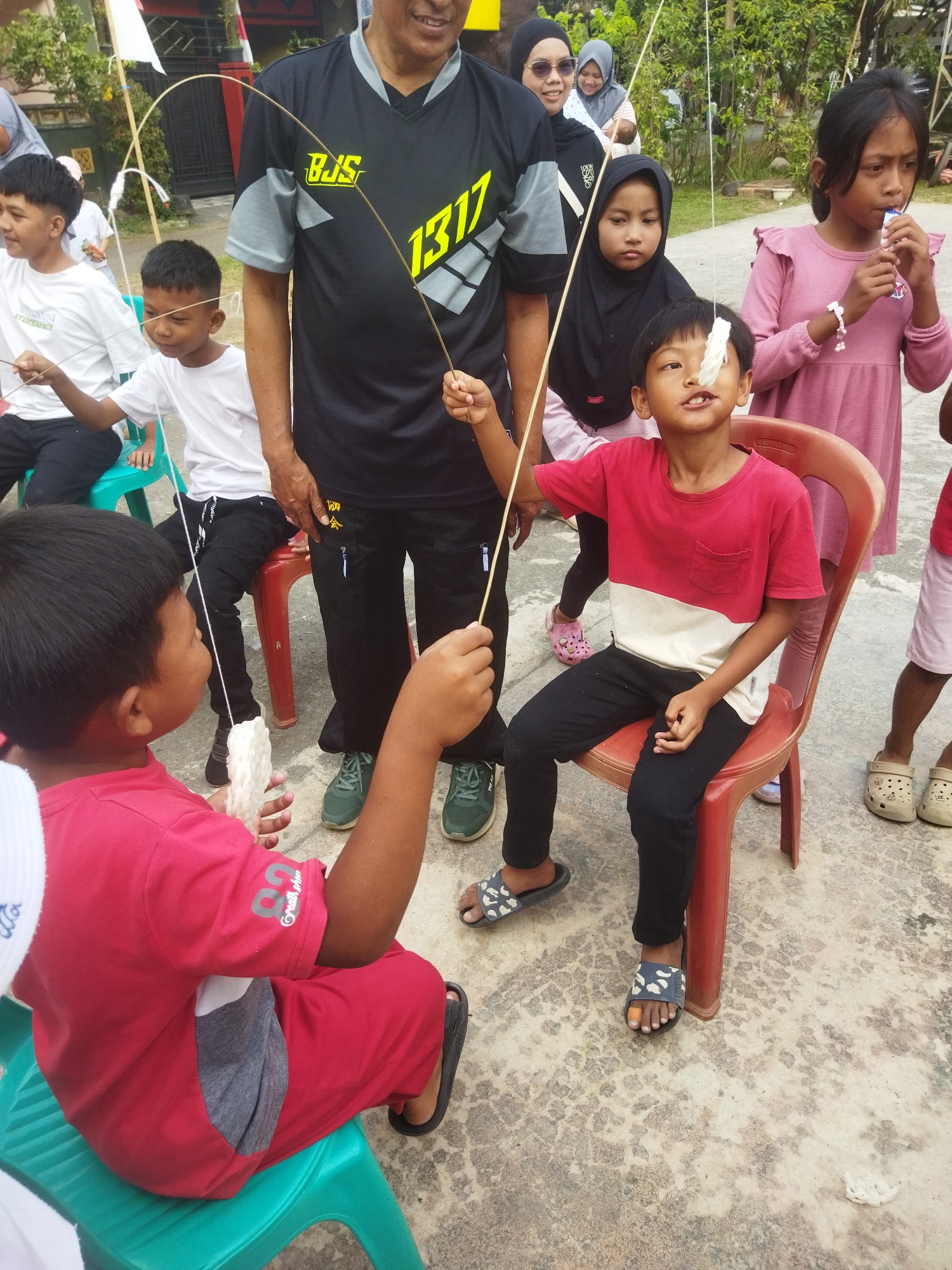 The image depicts an outdoor scene in the city of Banjar, Indonesia, where a group of people, primarily children and adults, are participating in what appears to be a game. In the foreground, a young boy in a red shirt is seated on a green chair, holding a piece of white, puffed rice-like food attached to a string. He is attempting to eat it without using his hands, as evidenced by his mouth being close to the food. To his right, another boy, also in a red and white shirt, is seated on a red plastic chair, looking up at a similar piece of food dangling from a string. He also seems to be engaged in the same eating challenge.
Behind them, an adult male wearing a black t-shirt with yellow lettering is standing, holding a string with food attached. Several other children and adults are visible in the background, observing the activity. The environment appears to be a paved area, possibly a yard or a community space, with some vegetation and structures in the distance. The overall atmosphere suggests a festive or recreational event.