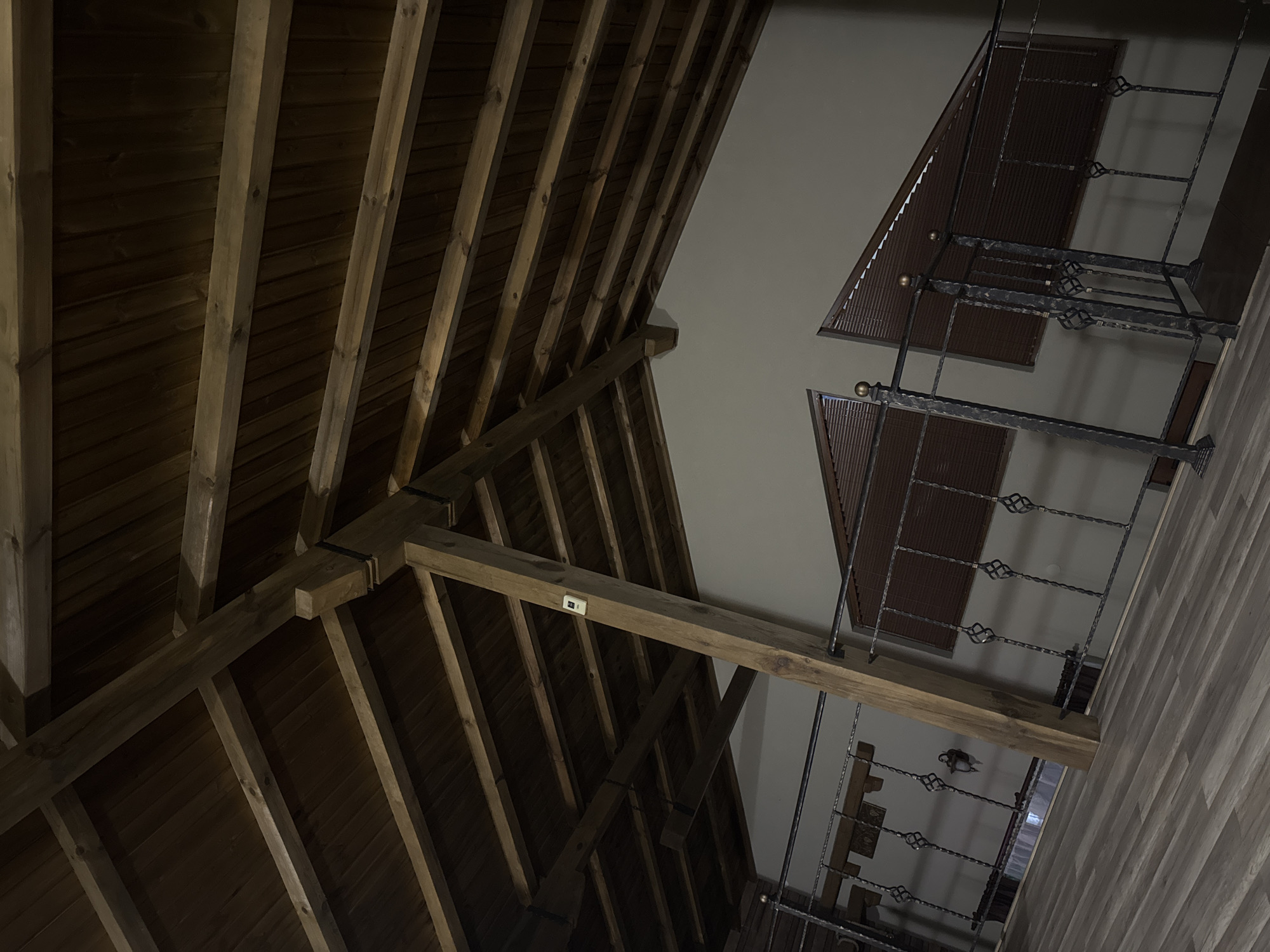 The image depicts an interior view of a building in Dolni Marian, Bulgaria, featuring exposed wooden roof beams and a section of a decorative wrought-iron railing and staircase. The roof structure consists of a series of parallel wooden beams supporting a wooden ceiling. The wrought-iron railing has a repeating pattern of decorative twists. The adjacent wall and ceiling are painted white. The floor is covered with light-colored, wood-patterned planks.
