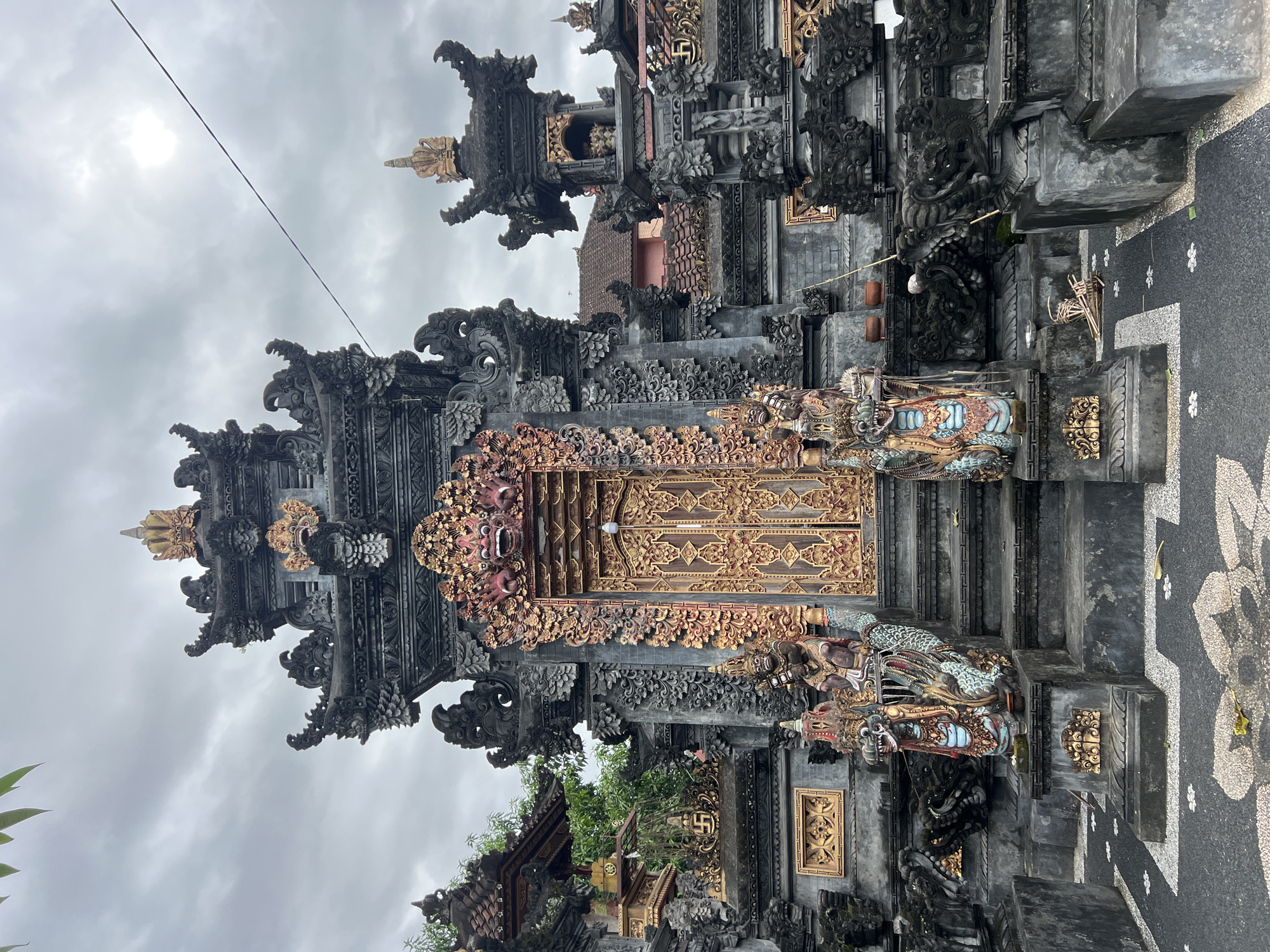 This image depicts the ornately decorated entrance to a Balinese temple. The entrance features a towering structure of dark gray stone, intricately carved with floral and mythical motifs. A pair of golden doors are flanked by multi-colored statues of mythical creatures. The surroundings are a blend of stone paths marked with floral patterns and parts of other temple structures, all under a cloudy sky. This is likely located in the city of Sukawati, Indonesia, a region known for traditional Balinese art and architecture.