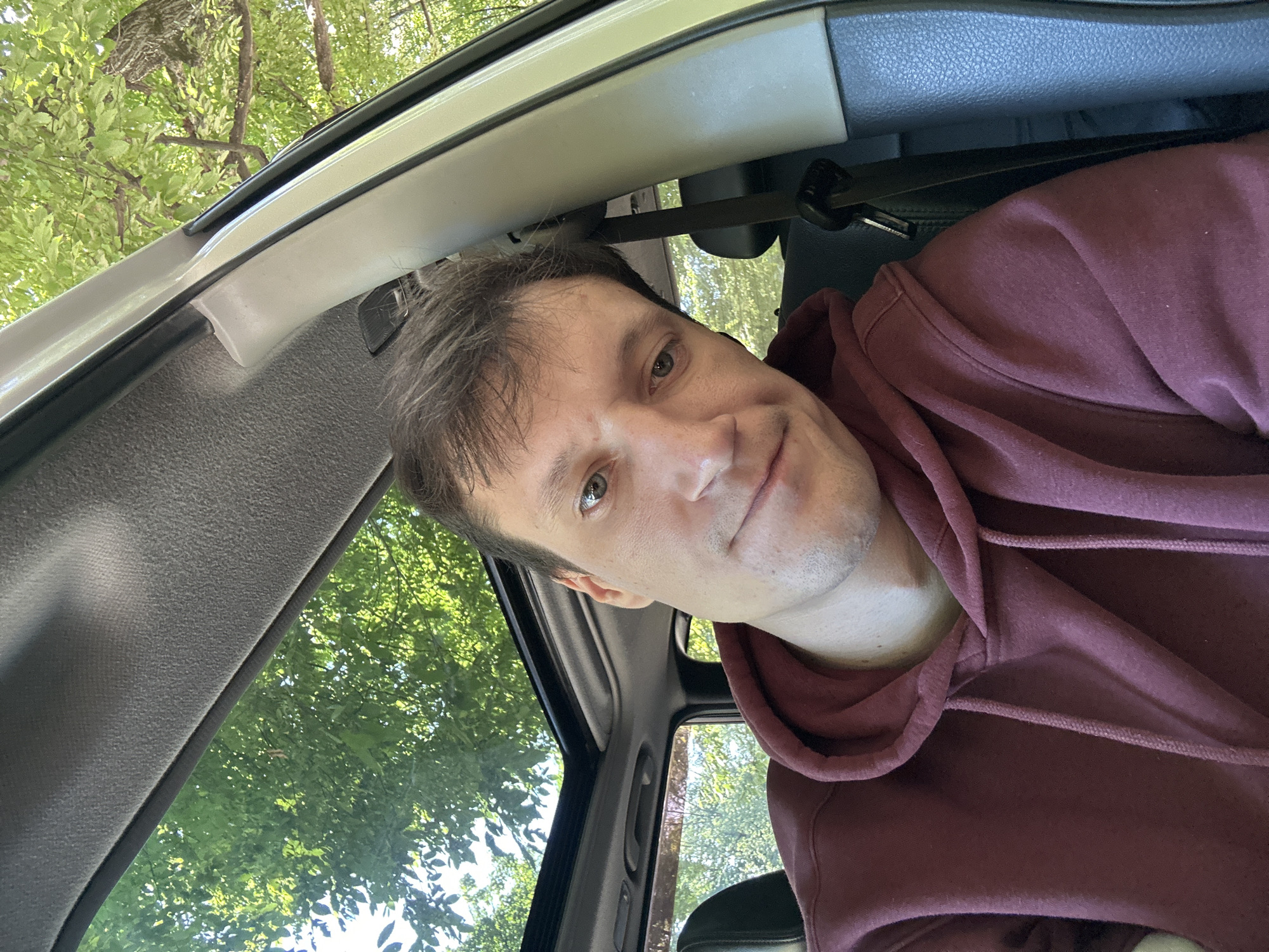 A man wearing a maroon hoodie is sitting inside a car with the sunroof open, looking at the camera with a slight smile. Green trees and foliage are visible through the sunroof and the car windows, indicating an outdoor setting, possibly in a wooded area. The interior of the car, including the headliner and a seatbelt, is also visible. The man's face and upper body are the main focus of the image. There is no visible indication of the city of Shatava, Ukraine.