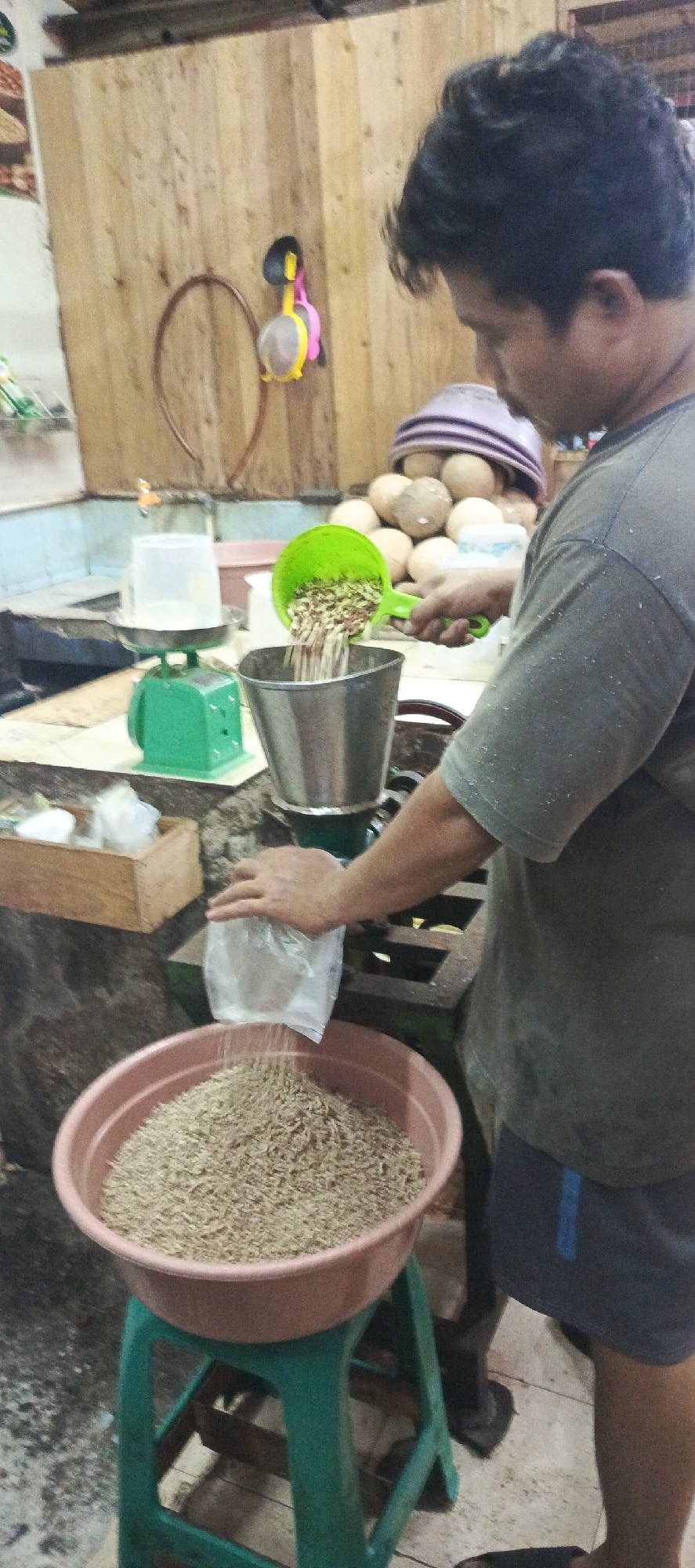 In an indoor setting, likely a market stall in Sukaresmi, Indonesia, a man is actively engaged in processing a granular substance. He is wearing a grey t-shirt and dark shorts. In his right hand, he holds a green scoop filled with what appears to be grains or seeds, which he is pouring into a silver metal container positioned on a dark apparatus. Simultaneously, with his left hand, he holds a clear plastic bag from which a finer, lighter-colored granular material is being poured into a large, pinkish-brown basin. This basin is elevated on a green plastic stool.
Behind the man, a wooden wall is visible, adorned with a circular object and a yellow and pink item hanging from a hook. In the background, several spherical objects, possibly coconuts or similar produce, are stacked, some covered with a purple cloth. To the left, a green weighing scale with a clear container on top is present, along with a wooden box containing small white packets. The floor appears to be tiled or concrete. The overall impression is of a food processing or packaging operation.